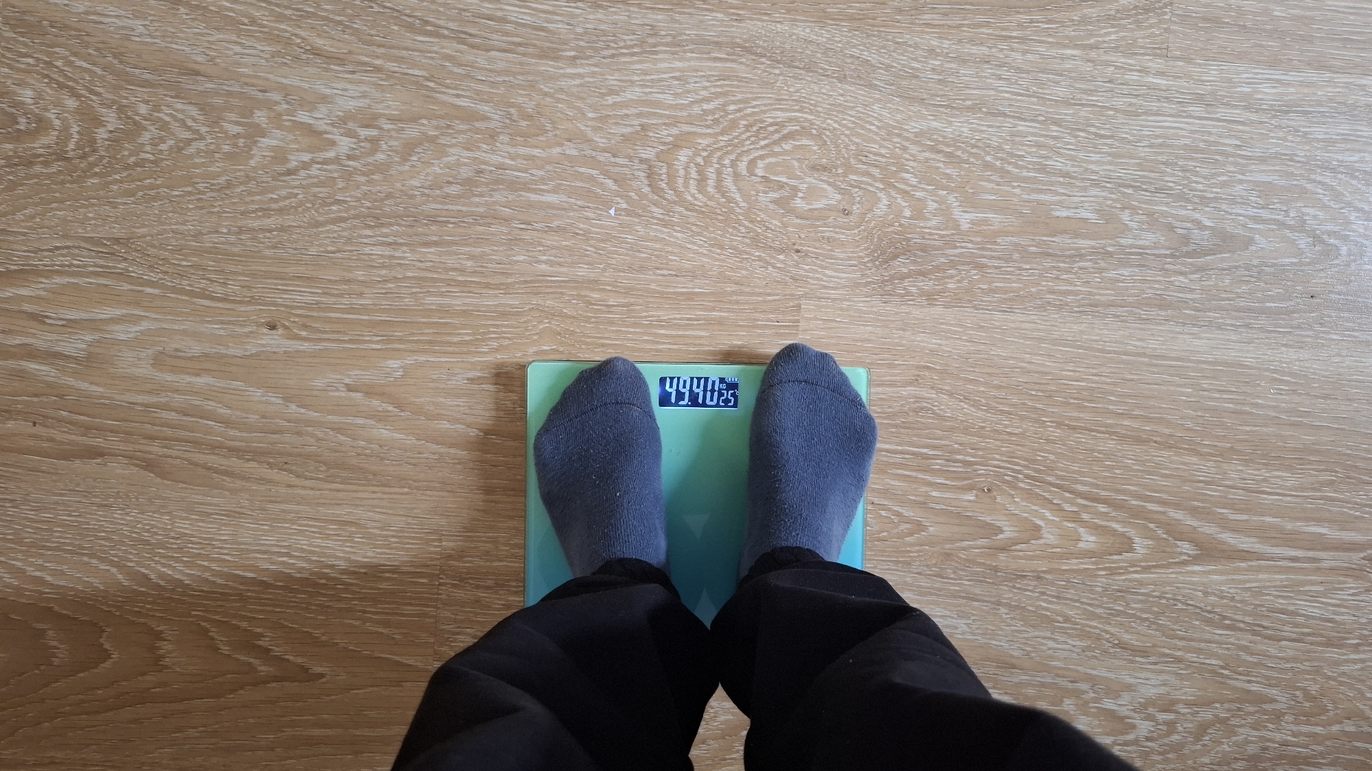 The image captures a first-person, downward view of a person standing on a digital bathroom scale. The person's feet, clad in dark gray socks, are positioned centrally on the light green or teal-colored scale. Dark trousers are visible around their lower legs.
The digital display on the scale clearly shows the numbers "49.40" in blue, followed by "KG" (kilograms) in smaller text, and an even smaller "25" below and to the right of "KG". This indicates a weight measurement of 49.40 kilograms, with the "25" potentially representing a body fat percentage or another body composition metric.
The scale rests on a light-colored wooden floor, which exhibits prominent grain patterns consistent with laminate or hardwood flooring. The lighting suggests an indoor setting. No other individuals, specific time of day, or environmental details are visible. The activity depicted is the act of a person weighing themselves.