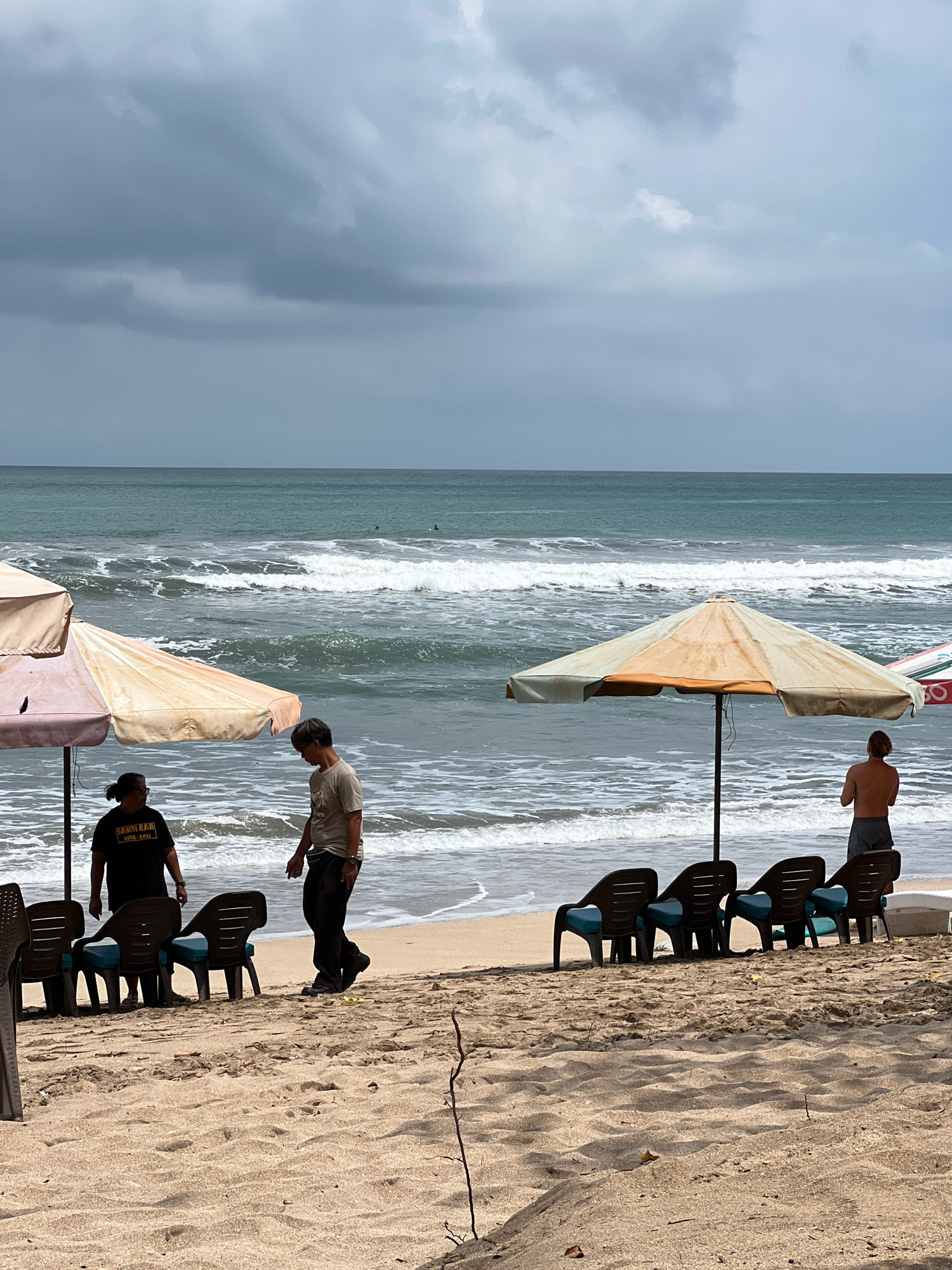 The image depicts a beach scene in Kuta, Indonesia, under a cloudy sky. Two beach umbrellas are set up on the sand, providing shade. In the foreground, several plastic lounge chairs are arranged facing the ocean. Two individuals are visible near the left umbrella. One person, wearing a black t-shirt with text, stands with their back to the camera, while another person in a light-colored t-shirt and dark pants stands facing them. Further to the right, a shirtless individual wearing dark shorts stands facing the ocean, with another umbrella and more chairs in the vicinity. The ocean has moderate waves, and a few distant figures can be seen in the water, possibly surfers. The sand appears light brown and textured, with some scattered debris. The overall atmosphere suggests a somewhat overcast day at the beach.