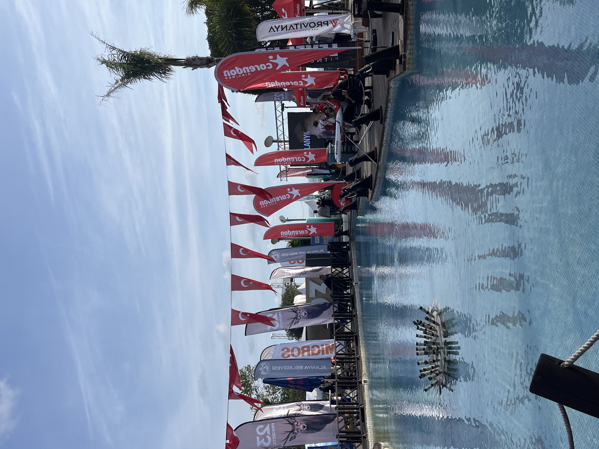 An outdoor event is observed at an aquatic venue in Alanya, Turkey. The scene is dominated by a large, light blue tiled pool, bordered by a dark wooden deck. A dark wooden railing with a white rope accents the immediate pool edge.
Within the pool, a floating decorative fountain element, composed of multiple dark cylindrical pipes arranged radially, is visible. Above the event area, numerous red and white Turkish national flags are strung horizontally. Along the wooden deck, a series of tall, vertical advertising banners are erected. These include red banners featuring the "Corendon" logo and white text, and grey/white banners depicting a deer head with antlers. The word "ALANYA" is visible on several banners and prominently displayed on a large LED screen in the background.
A white and red model airplane rests on a stand on the wooden deck. Several individuals are present on the deck, standing and seated. Palm trees and other green vegetation are situated behind the event structures, beneath a partly cloudy blue sky. Reflections of the banners and surrounding elements are visible on the pool's surface.