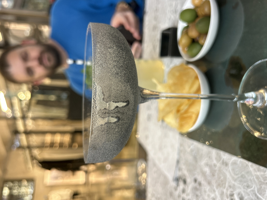 The scene depicts an indoor establishment in Milan, Italy. In the foreground, a clear stemmed coupe glass is centrally positioned, its bowl displaying a frosted, grey granular exterior with vertical clear streaks suggesting condensation. The glass contains a dark liquid. It rests on a reflective, dark teal surface. To the immediate left, a white oval bowl contains golden-yellow potato chips. To the right, another white oval bowl holds an assortment of green and brown olives. A small black rectangular dish is situated behind the olive bowl. The underlying table surface is light-colored with a subtle, mottled pattern.
In the blurred midground, a man with a beard is visible, wearing a blue long-sleeved shirt. His facial features are indistinct due to the shallow depth of field. His right hand is seen holding a dark object. A glass containing a yellow liquid is partially visible behind the frosted coupe glass. The background is heavily blurred, showing bokeh effects from multiple artificial light sources and indistinct shapes indicative of interior furnishings or racks within a commercial space.