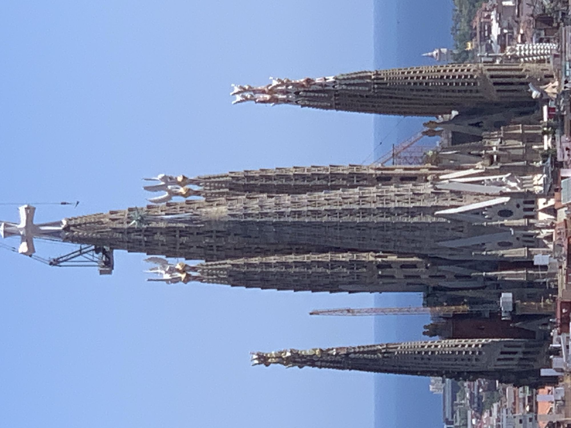 The image shows a close-up, eye-level view of the Sagrada Familia basilica in Barcelona, Spain. The basilica is under construction, indicated by a crane positioned near the top of one of its spires. The architecture is highly intricate and Gothic in style, with numerous spires, statues, and detailed carvings. The basilica is a dominant structure in the frame, set against a clear blue sky. Below the basilica, the city of Barcelona can be seen with its rooftops and some distant structures, including a lighthouse or tower. The sea is visible in the background, bordering the cityscape. The overall scene depicts a landmark under development, showcasing its impressive scale and architectural detail.