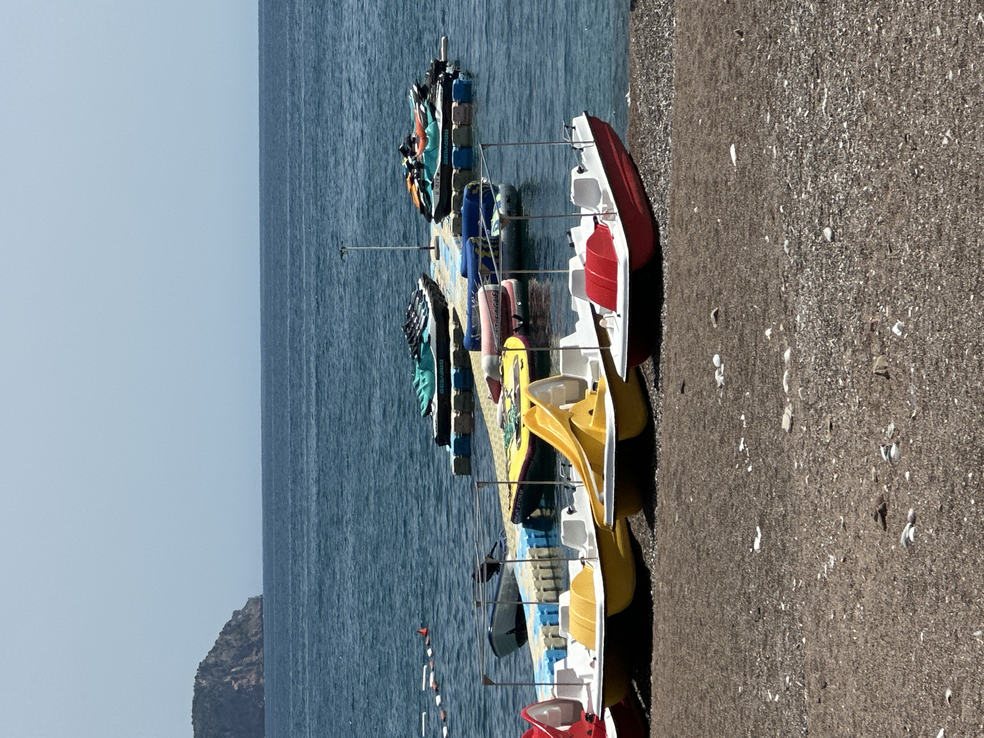 This is a photograph taken in Boreti, Montenegro, showcasing a beach scene with various watercraft. The foreground features a pebbled beach with several colorful pedal boats lined up. These boats are predominantly yellow and red, with white seating and canopies.
In the middle ground, a floating dock extends into the calm, blue water. Moored to this dock are several jet skis, some in green and white, others in orange and black. There are also a couple of inflatable tubes or rafts visible. A small, white motorboat is also tied to the dock.
The background is dominated by the expanse of the Adriatic Sea under a clear, pale blue sky. To the far left, a lush, green, rocky island or peninsula juts out of the water. The overall impression is a recreational area on a sunny day. There are no people visible in the frame.