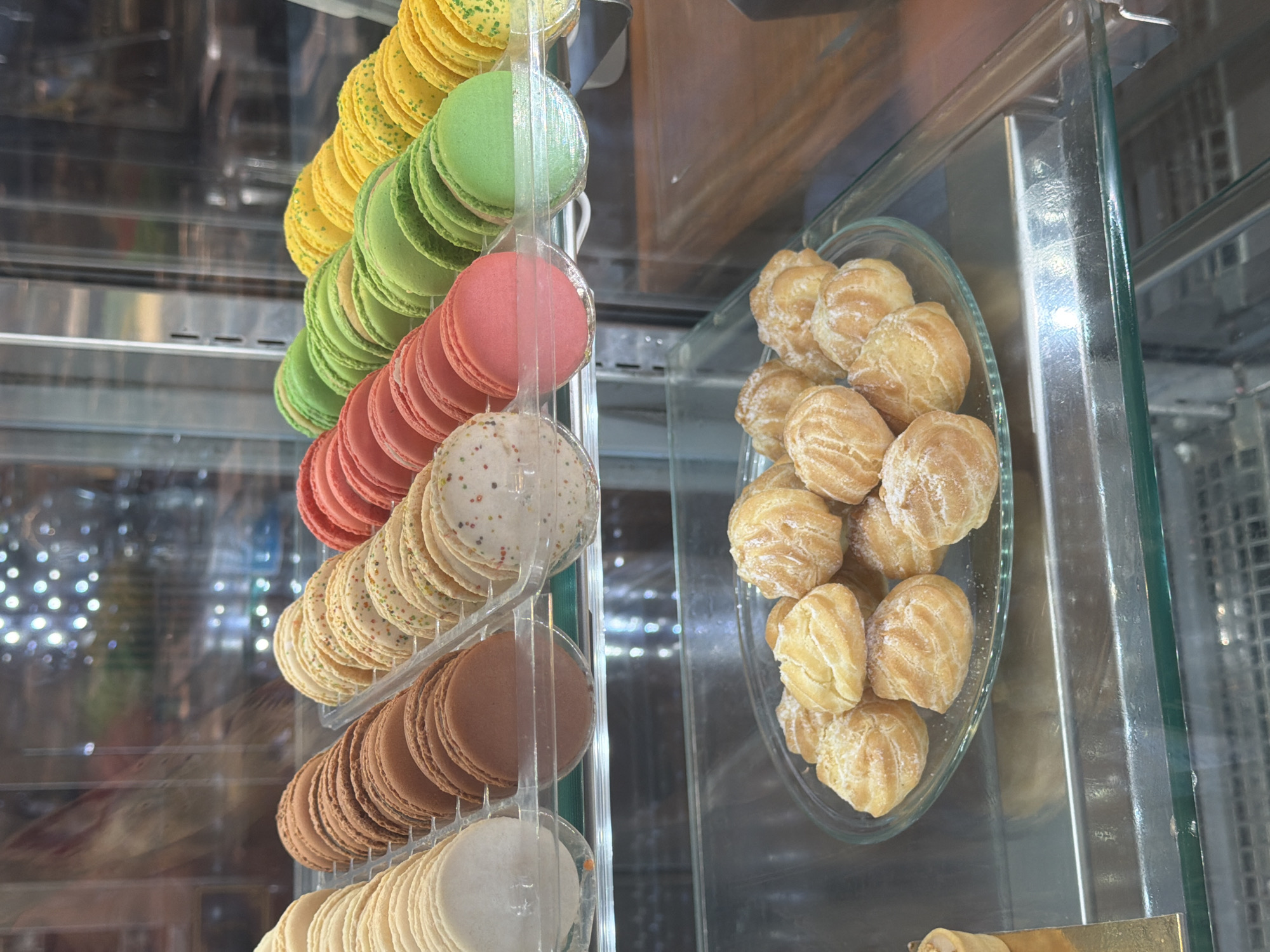 The image shows a display case, possibly in a bakery or pastry shop in Unknown, Unknown. A vertical rack holds rows of colorful macarons in shades of yellow, green, red, brown, and cream. Beside the macarons, a clear glass dish contains a collection of cream puffs dusted with powdered sugar. The display case is made of clear glass with metal trim.