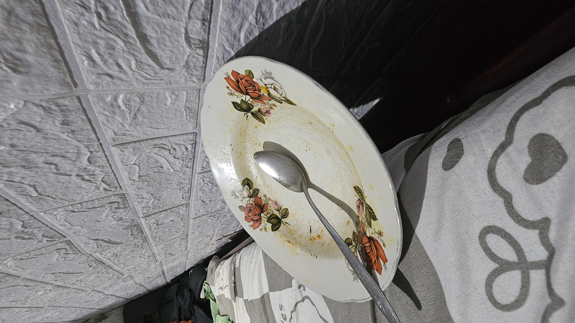 A dirty oval-shaped plate with a floral pattern of red and pink roses and green leaves sits on a patterned cloth. A metal spoon rests on the plate, which appears to have remnants of food. The plate and spoon are illuminated by a light source from the left, casting a shadow on a textured, brick-like wall in the background. The scene is likely indoors, potentially in a domestic setting within Bandung City, Indonesia. The overall impression is one of a used dish, perhaps after a meal.