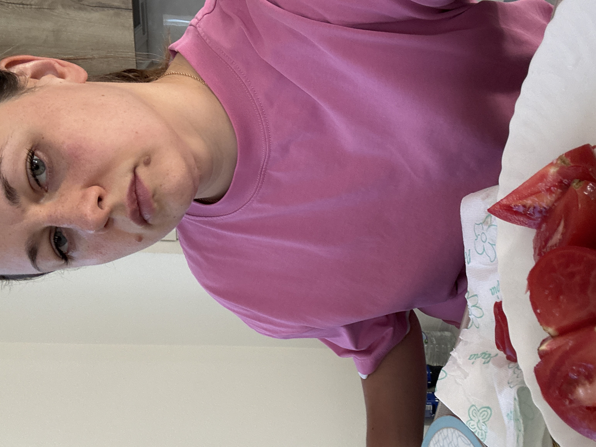 A medium shot captures a woman from the chest up, positioned in what appears to be a domestic setting. She is wearing a bright pink t-shirt. Her gaze is directed slightly upwards and to her right, with a neutral expression. The background is a plain, light-colored wall, with a hint of kitchen cabinetry visible on the right side. In the foreground, a white paper towel with a floral pattern is visible, alongside slices of red tomatoes, suggesting food preparation. There are no overt signs of distress or danger in the image. The location of Uyutnoye, Ukraine is not identifiable from the visual information provided in the image.