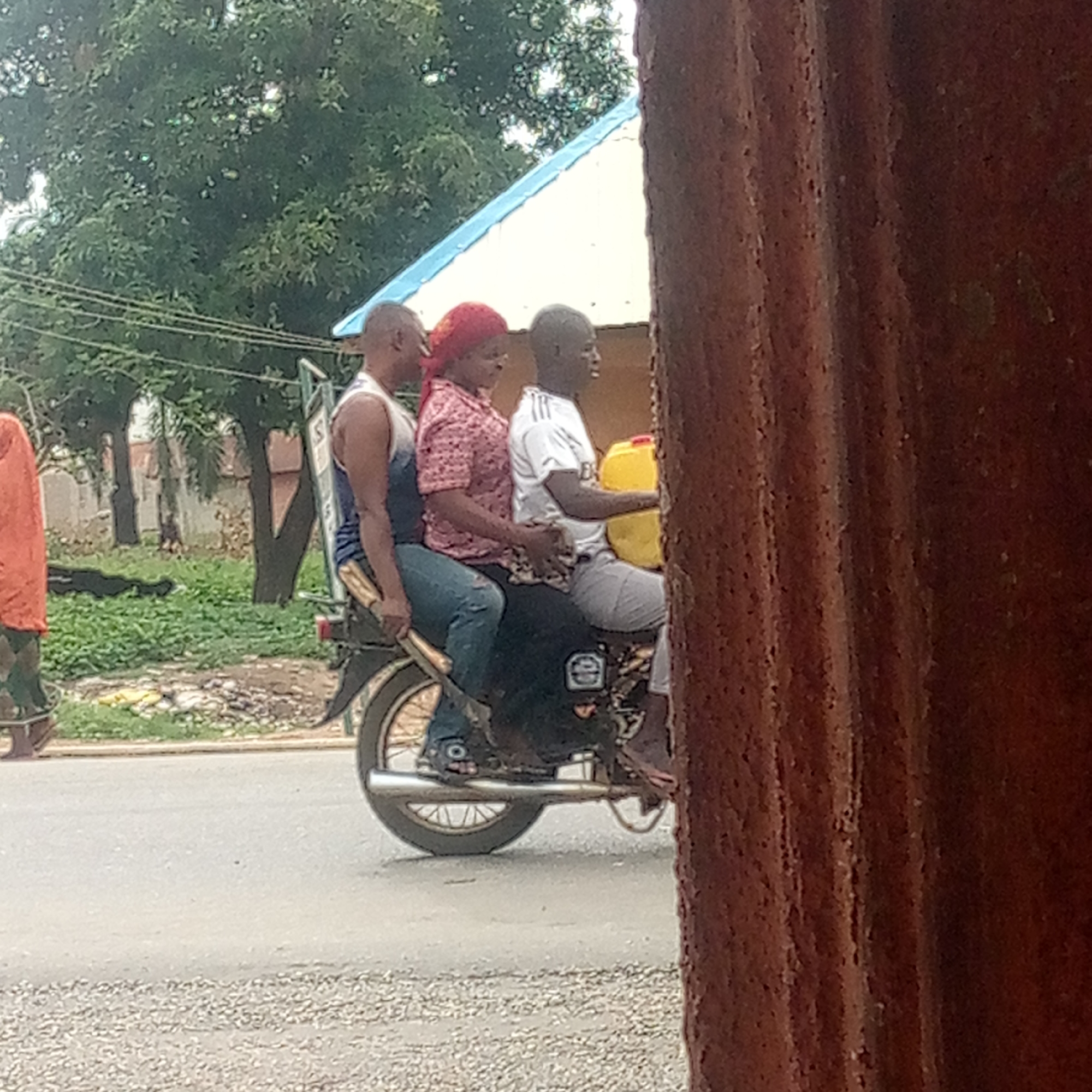Three individuals are riding on a motorcycle on a street in Mararaba, Nigeria. The individual in the front is operating the motorcycle, while holding a yellow plastic container. A woman with a red headscarf sits in the middle, and a man is positioned in the back. The motorcycle is moving on the asphalt street. In the background, there are trees and a building. A person is seen walking on the side of the road, and a textured brown structure is partially obstructing the right side of the view.
