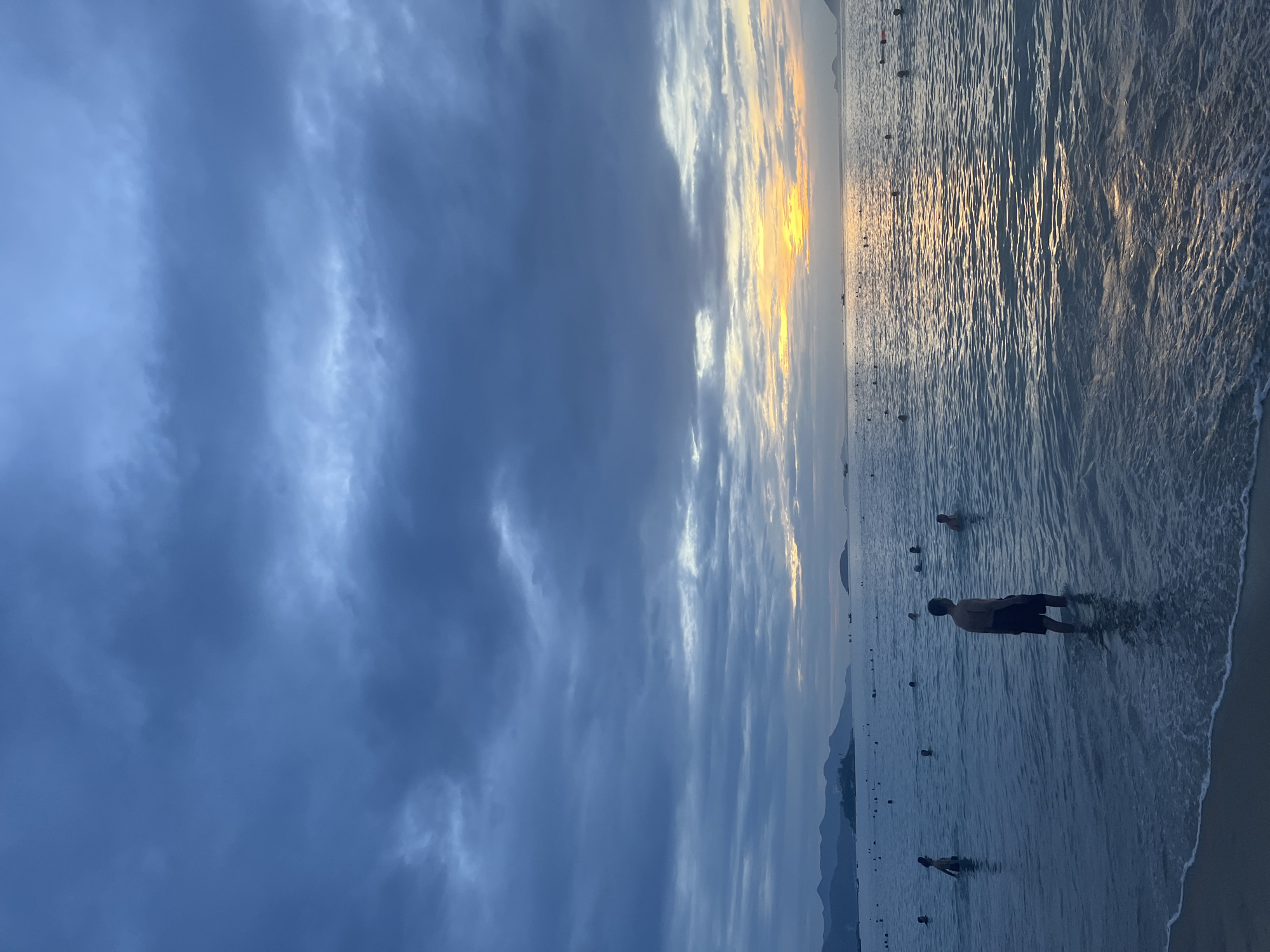 The image depicts a beach scene in Nha Trang, Vietnam, during twilight. The sky is filled with dramatic, dark blue clouds, but a sliver of golden sunset is visible on the horizon, reflecting on the calm sea. Several people are in the water, some wading and others appearing to swim. A man stands in the foreground, facing away from the camera, with the sea reaching his knees. In the distance, a silhouette of mountains lines the coast. The overall atmosphere is serene and tranquil.