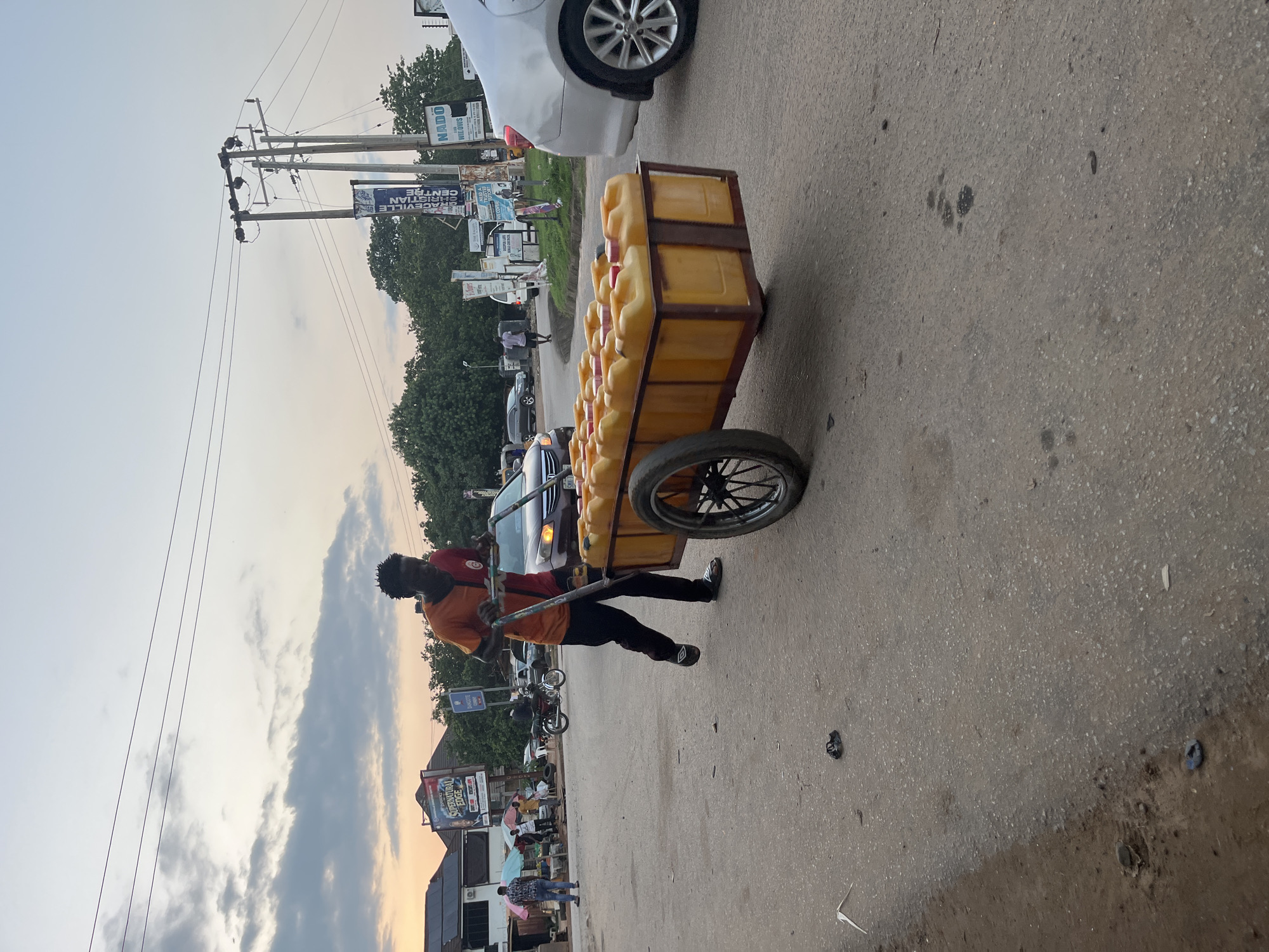A man is pushing a cart laden with yellow jerrycans of water down a street in Abuja, Nigeria. The man is wearing an orange and red shirt and dark pants. The cart has two wheels and is pulled by handles. The street is paved with asphalt and appears to be a moderately busy thoroughfare with other vehicles and people visible in the background. There are buildings and trees lining the street, and power lines are visible overhead. The sky is partly cloudy with the sun setting or rising. The overall impression is one of everyday commerce and street life in an urban environment.