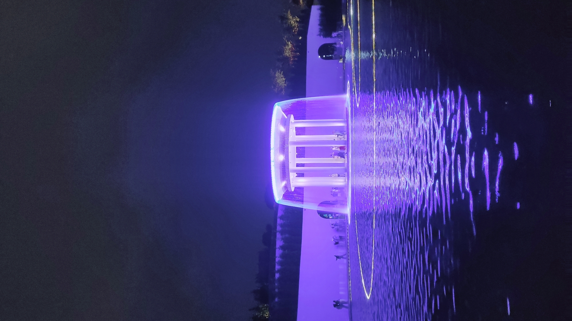The image depicts a modern fountain structure illuminated with purple lights at night. The fountain is circular with a series of tall, white columns in the center, supporting a circular canopy from which water cascades downwards, creating a curtain-like effect. The water falls into a shallow pool that reflects the purple illumination and features a circular pattern of yellow lights on its surface. In the background, a long, white wall also glows with purple light, and a row of trees is visible beyond it. Several people are standing around the fountain, some on the walkway bordering the pool and others within the illuminated area of the fountain itself. The overall scene is one of a public space designed for nighttime viewing and entertainment, with a focus on light and water features. The specific location of this fountain in Krasnodar, Russia, is not provided by the image itself.