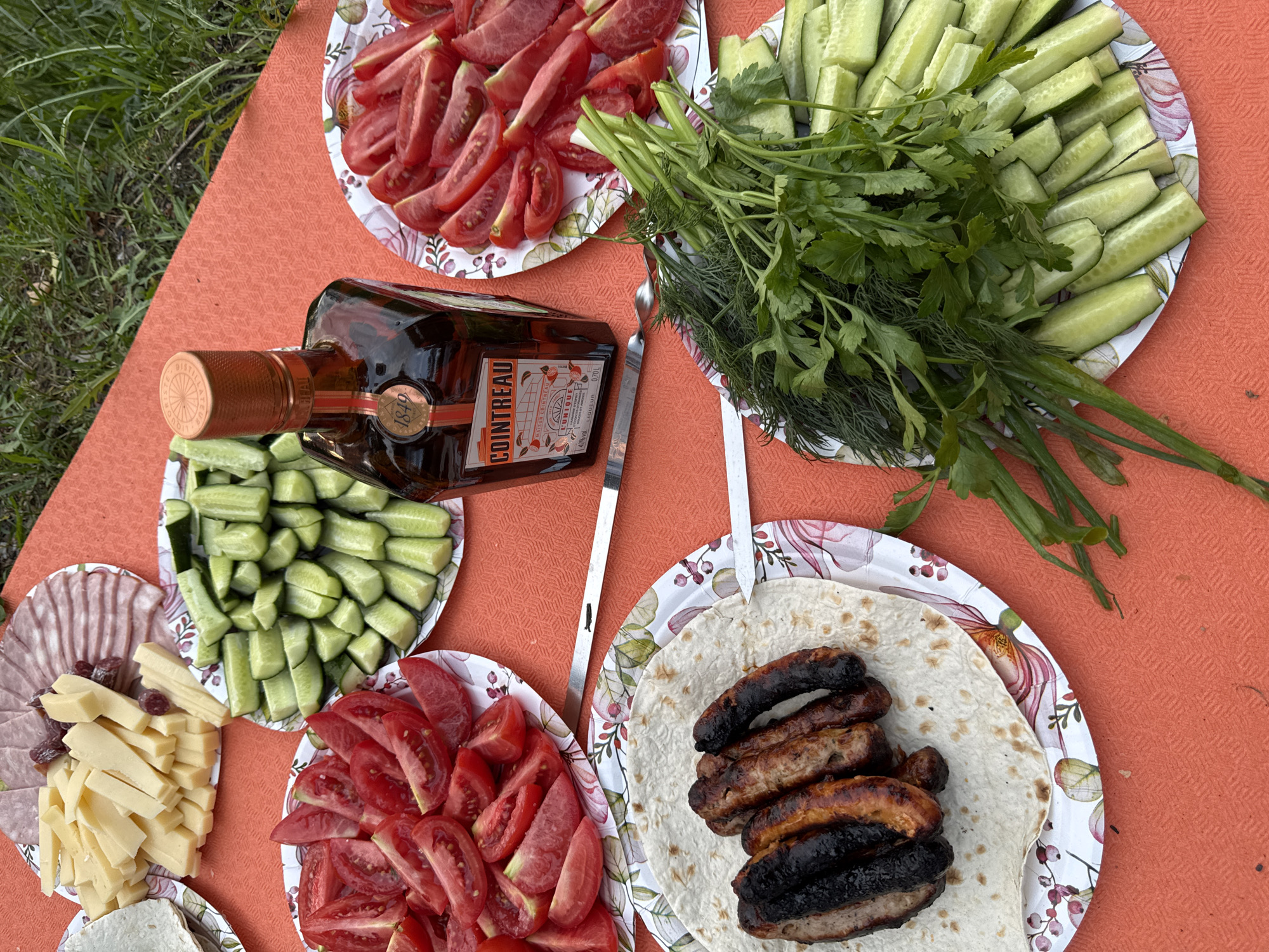 A top-down, medium shot captures a picnic spread on an orange, textured tablecloth outdoors, with grassy terrain visible in the background. The arrangement features several plates of food and a bottle of Cointreau liqueur. On the left, a plate holds slices of cheese and cured meats. Adjacent to it, a plate is piled high with sliced tomatoes. Further down, a flatbread is topped with several grilled sausages, some appearing burnt. To the right of the flatbread, a plate displays chopped cucumbers. A large bunch of fresh herbs, including dill and parsley, rests on top of another plate of sliced cucumbers and green onions. A silver skewer lies across the flatbread and the plate of herbs, connecting the two. The Cointreau bottle, with its distinctive orange cap and label, stands centrally in the upper portion of the frame, positioned between the plates of vegetables and cheese. The scene appears to be set up for a casual outdoor gathering.