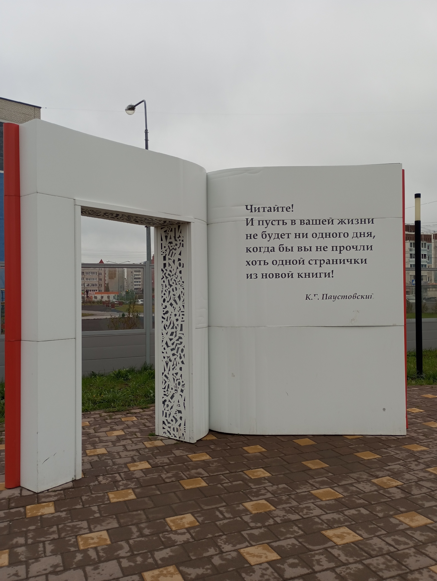 Here's a detailed description of the image:
The photo presents a large, walk-through book-shaped structure in Kamensk-Uralsky, Russia. The "book" is primarily white, with red accents on the spine. The open "pages" display text in Russian:
"Читайте! И пусть в вашей жизни не будет ни одного дня, когда бы вы не прочли хоть одной странички из новой книги! К.Г. Паустовский" which translates to "Read! And may there not be a single day in your life when you have not read at least one page from a new book! K.G. Paustovsky."
One "page" serves as an opening or doorway, framed with white material containing decorative, abstract cut-out patterns.
The book sits on a paved area of brown and tan interlocking square stones. Behind the book, through the open "doorway," one can see a grassy area and a cityscape with apartment buildings under a cloudy sky. To the left of the book, there's part of a building with red and gray panels. There's also a streetlamp behind it. To the right of the book, there is a pole.