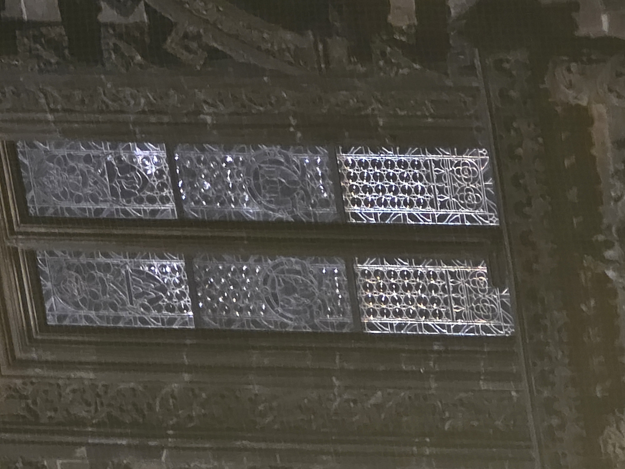 The image captures a detailed section of an ornate architectural element, likely part of a building's facade or interior wall. No people or specific activities are present. The primary subjects are two horizontal rows of decorative window-like grilles or panels, set within a larger, intricately carved structure.
The scene is dimly lit, with strong, bright reflections gleaming off the surfaces of the grilles, suggesting either an internal light source or sunlight striking the polished elements. The overall color scheme is dark and monochromatic, dominated by shades of grey, brown, and black, which accentuates the reflective highlights.
Each horizontal row of panels displays varying intricate patterns. The leftmost panels in both rows feature organic, flowing designs that resemble intertwined vines or stylized floral motifs. Adjacent panels, especially visible in the top row, continue with similar complex, organic, and perhaps symmetrical patterns. Towards the right side of both rows, the panels transition to more geometric designs, characterized by a repeating lattice of interlocking circles or oval shapes, creating a mesh-like appearance.
The area surrounding these grilles, both above and below, is composed of dark, heavily carved material, possibly stone or plasterwork. This material is adorned with elaborate relief carvings and moldings that complement the patterns in the grilles. The carvings appear abstract and decorative rather than depicting specific figures or scenes. The image is framed from a slightly upward perspective, focusing on these detailed architectural embellishments. No visible text is present in the image.