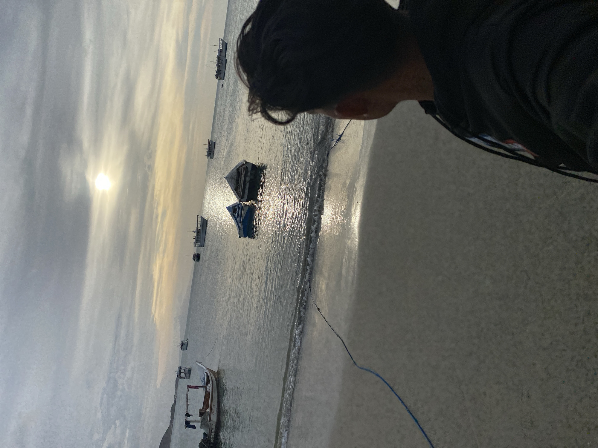 The image displays a coastal scene in Juan Griego, Venezuela, during what appears to be late afternoon or early evening. The foreground consists of light-colored sand, with a blue rope or hose extending across it towards the water. Gentle waves break on the sandy shoreline. A person, viewed from behind, occupies the lower right portion of the image, observing the water. This individual has dark hair and wears a dark hooded garment.
The midground and background feature a calm body of water populated by multiple boats. On the left side, closer to the shore, is a light-colored boat, possibly a fishing vessel, with an outboard motor mounted on the stern. A structure with fabric or clothing draped over it is visible above its stern. Text "PASSELITE" and "MARBELLA" is legible on its hull. Further out in the water, two smaller, partially submerged or derelict boats, one predominantly blue and the other white-grey, are centrally positioned. Several additional, larger vessels with dark hulls are scattered across the bay.
The sky is overcast with horizontal cloud formations. The sun is visible through a break in the clouds, creating a bright spot in the upper midground and casting reflections on the water's surface. A distant landmass, consistent with hills or mountains, is visible along the left horizon.