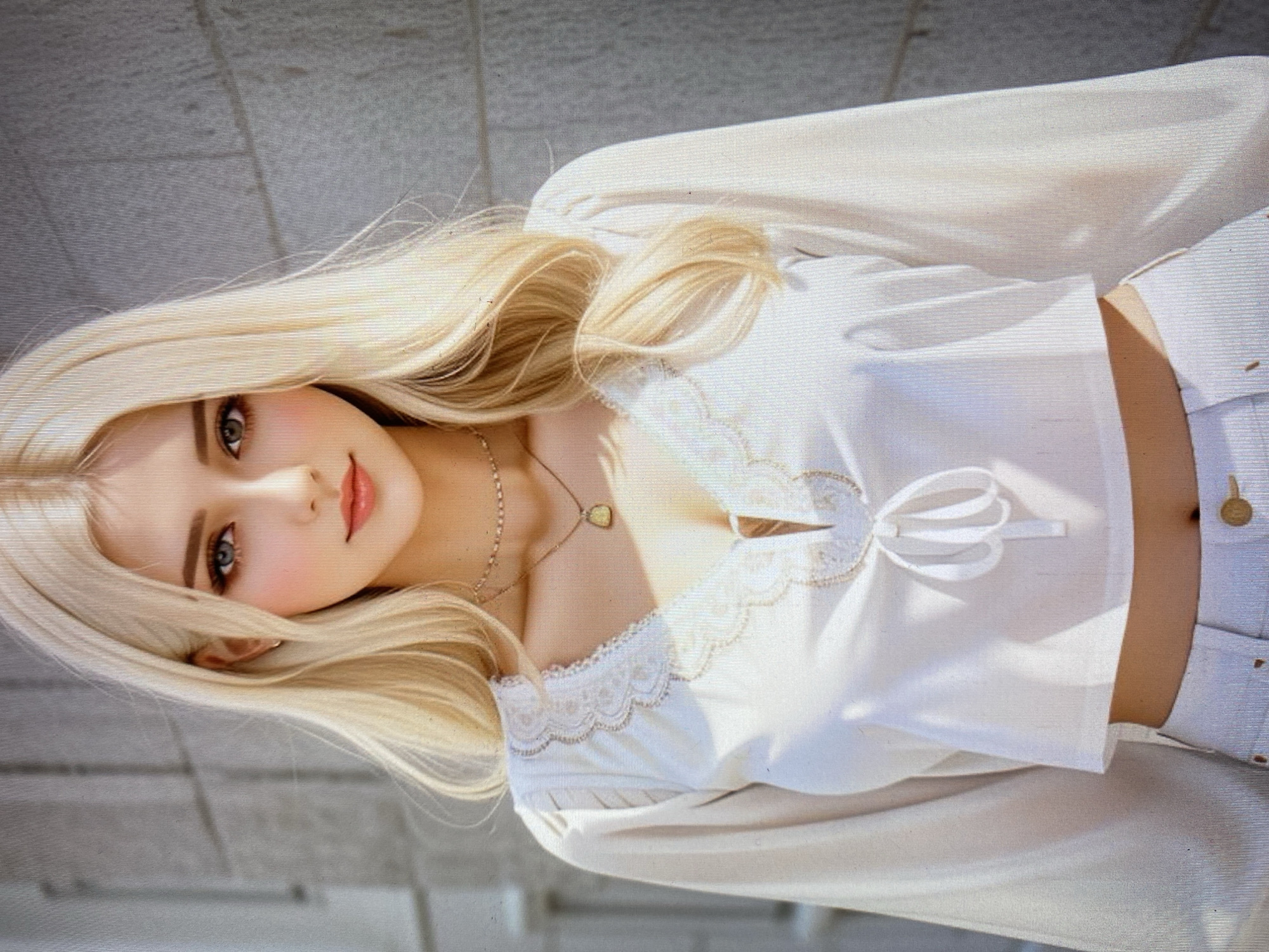 A medium shot shows a young woman with long, blonde hair and striking blue eyes. She is wearing a white, cropped blouse with intricate lace detailing around the neckline and cuffs. The blouse features a tie closure at the front. She is also wearing light-colored pants, with a button visible at the waist. The background consists of a rough, textured wall, suggesting an outdoor urban setting.