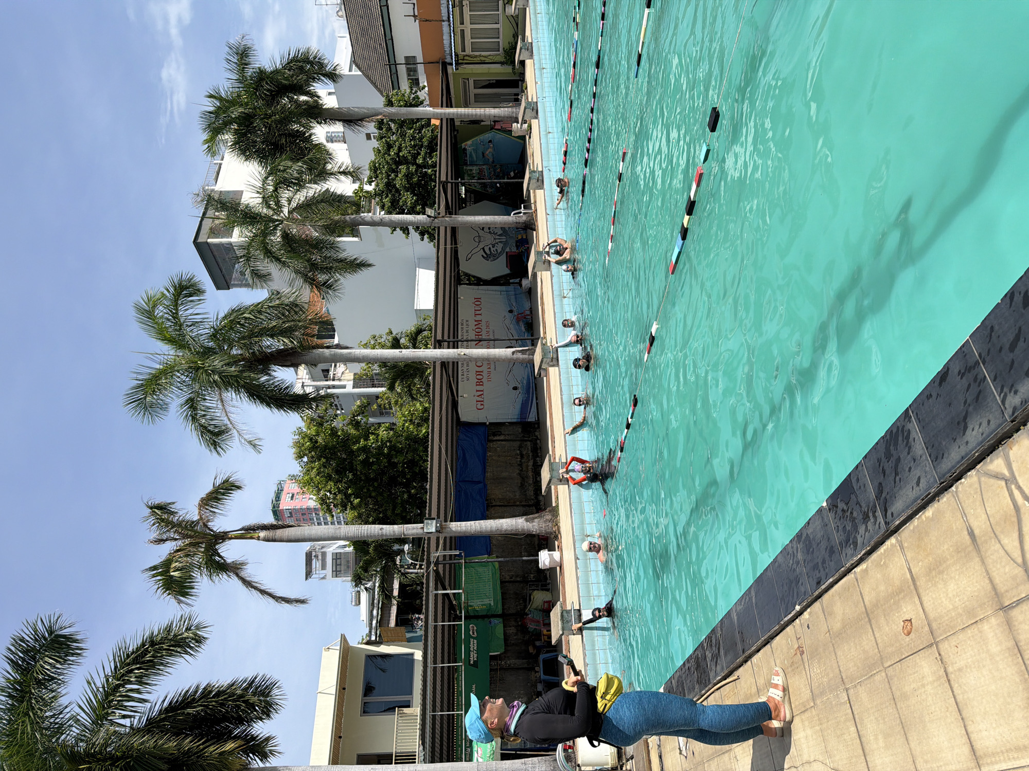In Nha Trang Ward, Vietnam, a swimming lesson is taking place at an outdoor swimming pool. The pool is rectangular, with lane ropes dividing it. Several participants, appearing to be children and adults, are in the water, some wearing swimming caps and goggles. A female instructor, wearing a black long-sleeved shirt, blue leggings, and a blue cap, stands on the pool deck, facing the swimmers and holding a yellow bag and a whistle. Palm trees surround the pool area, and buildings are visible in the background. The sky is clear and blue, indicating a sunny day.