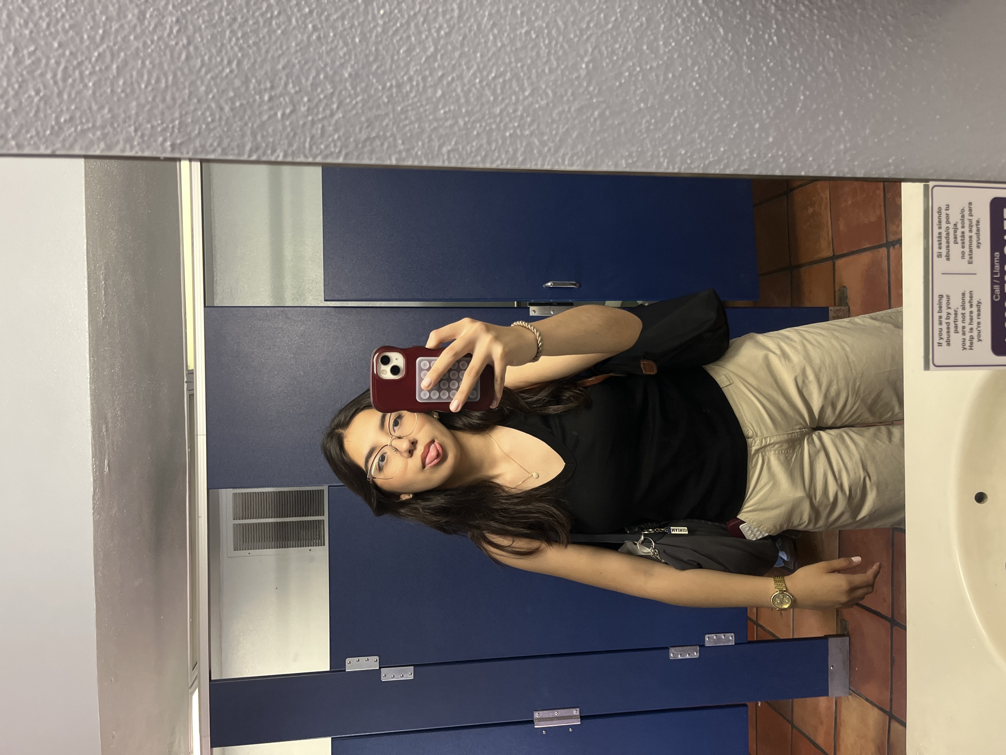 The image is a selfie taken in a bathroom, likely a public restroom, in the city of San Antonio, United States of America. A young woman with brown hair and glasses is visible, holding a smartphone with a maroon case. She is wearing a black top, khaki pants, and a gold watch. A black shoulder bag is slung across her body. The background features blue bathroom stalls, a white sink, and brown tiled flooring. A sign in English and Spanish with a domestic abuse hotline number is visible on the right side of the frame. An air vent is visible in the background.