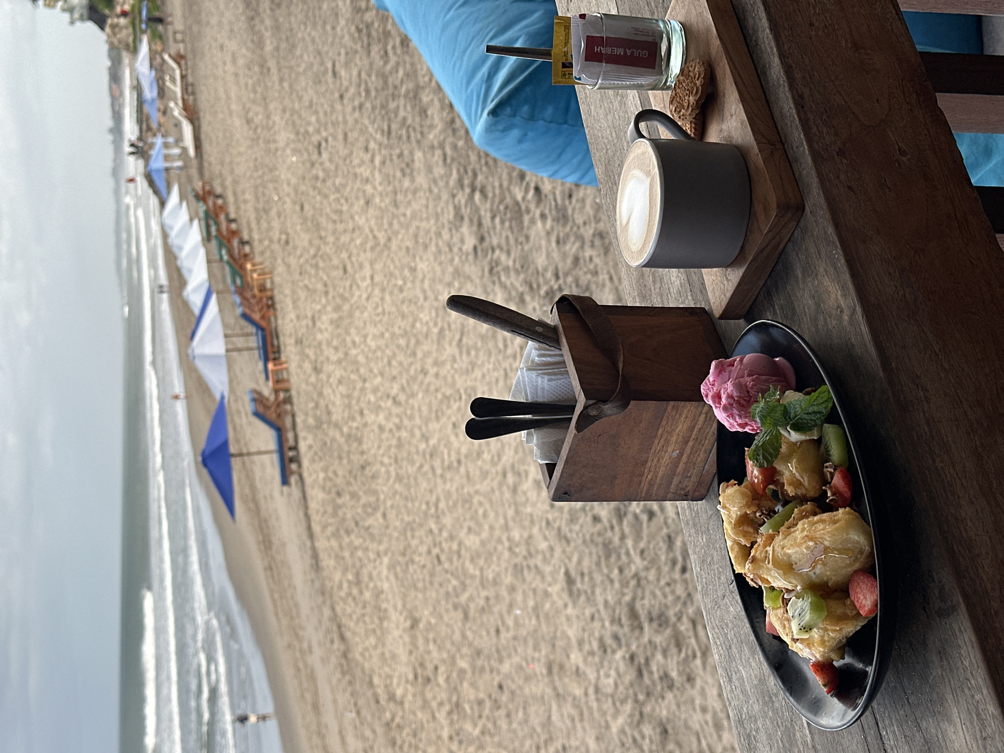 The image depicts a serene beach scene in North Kuta, Indonesia, likely in the early morning or late afternoon given the diffused lighting. In the foreground, a wooden table serves as a platform for a breakfast or dessert spread. A dark plate holds several pieces of fried pastry, generously drizzled with honey and topped with fresh strawberries, kiwi slices, and mint leaves. Adjacent to the pastry is a scoop of vibrant pink ice cream. To the right of the plate, a grey mug contains latte art, a heart shape formed by the milk foam, suggesting it's a cappuccino or similar coffee drink. Next to the mug, on a small wooden board, are two biscuits and a glass containing sugar packets. A wooden box filled with cutlery and napkins sits behind the plate.
The background transitions to a sandy beach with rows of empty lounge chairs and blue and white umbrellas, indicating a resort or public beach area. The ocean stretches out to the horizon with gentle waves rolling onto the shore. A few distant figures can be seen walking along the waterline, suggesting a quiet, uncrowded atmosphere. The sky is overcast, contributing to the soft, diffused light. The overall ambiance is one of relaxation and leisurely enjoyment of a meal by the sea.