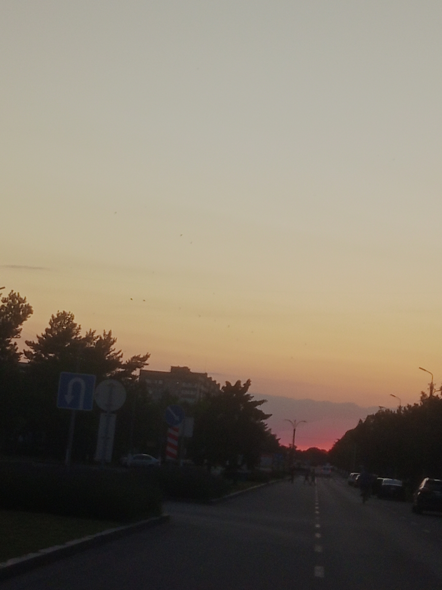 The image captures a road in Horishni Plavni, Ukraine, during sunset. The sky displays a gradient of pale yellow to orange and pink hues. On the left side of the road, there are trees and a building complex in the background. Road signs, including a U-turn sign, are visible near the trees. The road itself has dashed white lines marking lanes and is flanked by trees on both sides. Several cars are parked or driving along the road, and a few figures are present on or near the road. The overall impression is a suburban street scene at dusk.