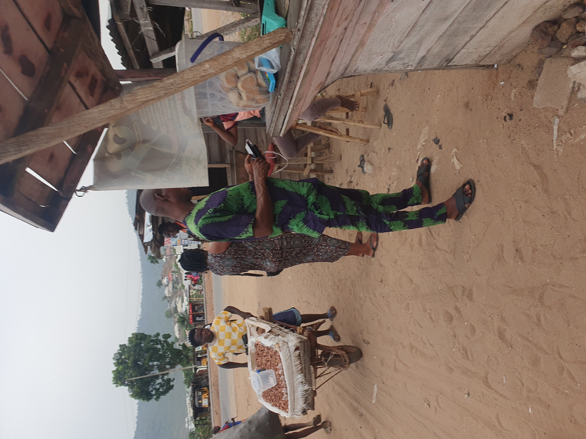 An outdoor scene in Lokoja, Nigeria, shows a bustling roadside market area under an overcast sky. The ground is predominantly light brown sand with visible footprints and scattered debris.
In the immediate foreground, a man stands wearing a green and purple patterned traditional outfit and black sandals. He is bald and looking down at a mobile phone he holds in his hands. Behind him, a woman is seen wearing a dark, patterned dress and sandals.
To their left, a young man, dressed in a yellow and white checkered shirt, blue shorts, and blue sandals, pushes a rustic handcart (or wheelbarrow). The cart is laden with a large, white sack filled with what appear to be roasted peanuts or groundnuts. A transparent plastic jug, possibly containing water, sits atop the sack.
The background reveals a local road, with distant views of buildings, scattered trees, and undulating hills, indicating a semi-urban or rural landscape.
Adjacent to the main figures is a simple wooden stall or shop structure, constructed from planks and timber. Inside the stall, a transparent plastic container holds several bread rolls or buns, suggesting a bakery or snack vendor. Another person's legs are visible seated behind the counter of this stall. A blue electronic device, possibly a payment terminal or small radio, is also visible on the stall's counter.
The overall atmosphere suggests a typical daily market activity, with people engaged in commerce or passing through. No specific text is clearly legible on any signs or objects.