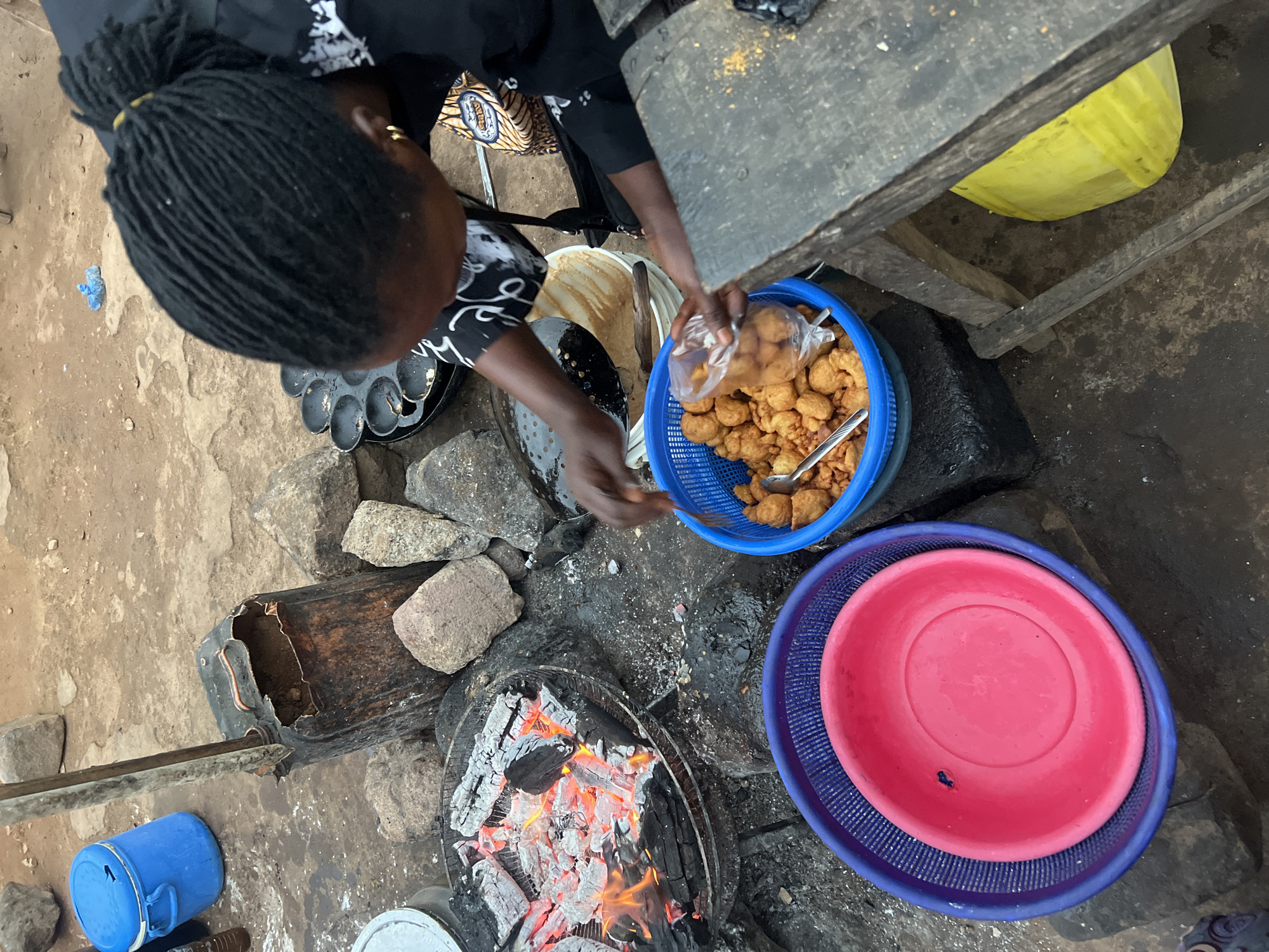 This overhead image depicts a person engaged in outdoor food preparation and packaging, likely a street vendor in Gura Top, Nigeria. The person, whose head and hands are visible, has dark braided hair pulled back. They are seen bagging freshly fried, golden-brown fritters, commonly known as puff-puff or similar dough balls, from a blue plastic basket into a clear plastic bag, indicating a transaction or preparation for sale. A silver spoon rests in the basket with the fritters.
The cooking setup is informal and rustic, situated on dirt and stone ground. A charcoal fire with glowing embers is central to the scene, surrounded by rocks that appear to support a cooking vessel (though not fully visible). Nearby, a pan with multiple circular indentations, possibly used for frying specific dough items, is present. A large white bowl containing a light-colored batter or dough suggests ongoing food preparation.
Other objects include various plastic containers: a blue plastic bucket on the ground, a yellow plastic bucket tucked underneath a rough wooden table, and a stack of a red plastic bowl inside a blue plastic strainer/colander. A makeshift metal container and a long wooden stick are also visible near the fire. The overall scene captures a typical bustling, informal food stall environment during the daytime, consistent with local market activities. There is no visible text in the image.