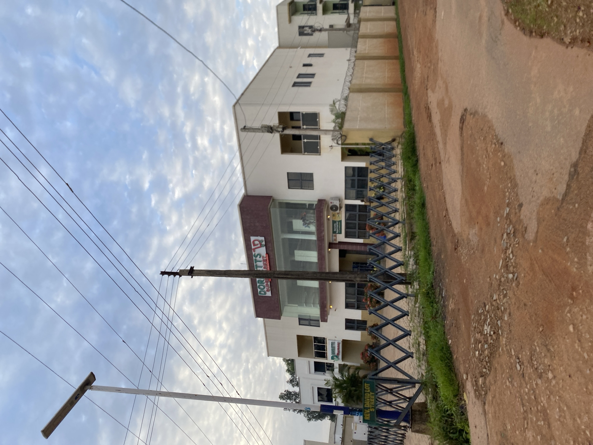 The image shows a street scene in Bar Jirgi Summa, Nigeria. The focus is on a multi-story building with a burgundy and white facade. The building appears to be a commercial establishment, with a sign reading "DORITTS SUPERMARKET" on the upper floor, and another sign that is partially visible lower down. There is also a sign for "REV. DAVID LOT STREET" visible in the lower right corner. The building is bordered by a black metal fence with a diamond pattern. Adjacent to the building is a dirt road with patches of green grass. Power lines crisscross the sky, which is partly cloudy. The lighting suggests it is daytime, possibly late afternoon or early morning due to the soft light. No people are clearly visible, but there are signs of human activity, such as the building itself and the road. The overall impression is a typical urban or semi-urban setting in Nigeria.