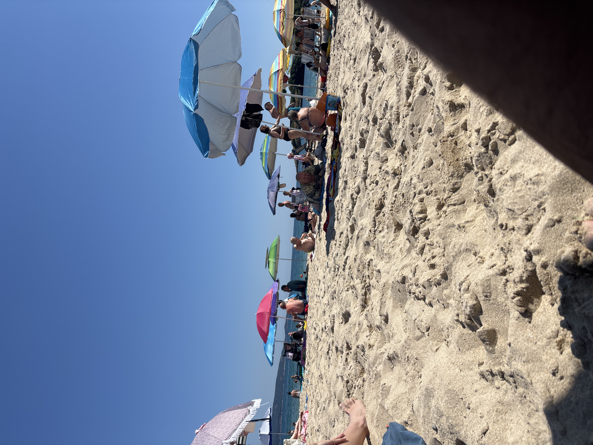 This is a photograph of a beach scene in Kiten, Bulgaria. The image is taken from a low angle on the sand, looking towards the sea. The beach is populated with many people, most of whom are seated or lying under colorful beach umbrellas. The sea is visible in the background, with a clear blue sky above. Several beach umbrellas are spread across the sand, in various colors including pink, blue, green, and striped patterns. Some individuals are partially visible in the foreground and mid-ground, some in swimwear. The sand appears to be light brown and textured, with footprints and small dunes. The water is calm and appears to be shallow near the shore. In the distance, a hazy coastline with some greenery can be seen. The overall impression is a sunny, crowded day at the beach.