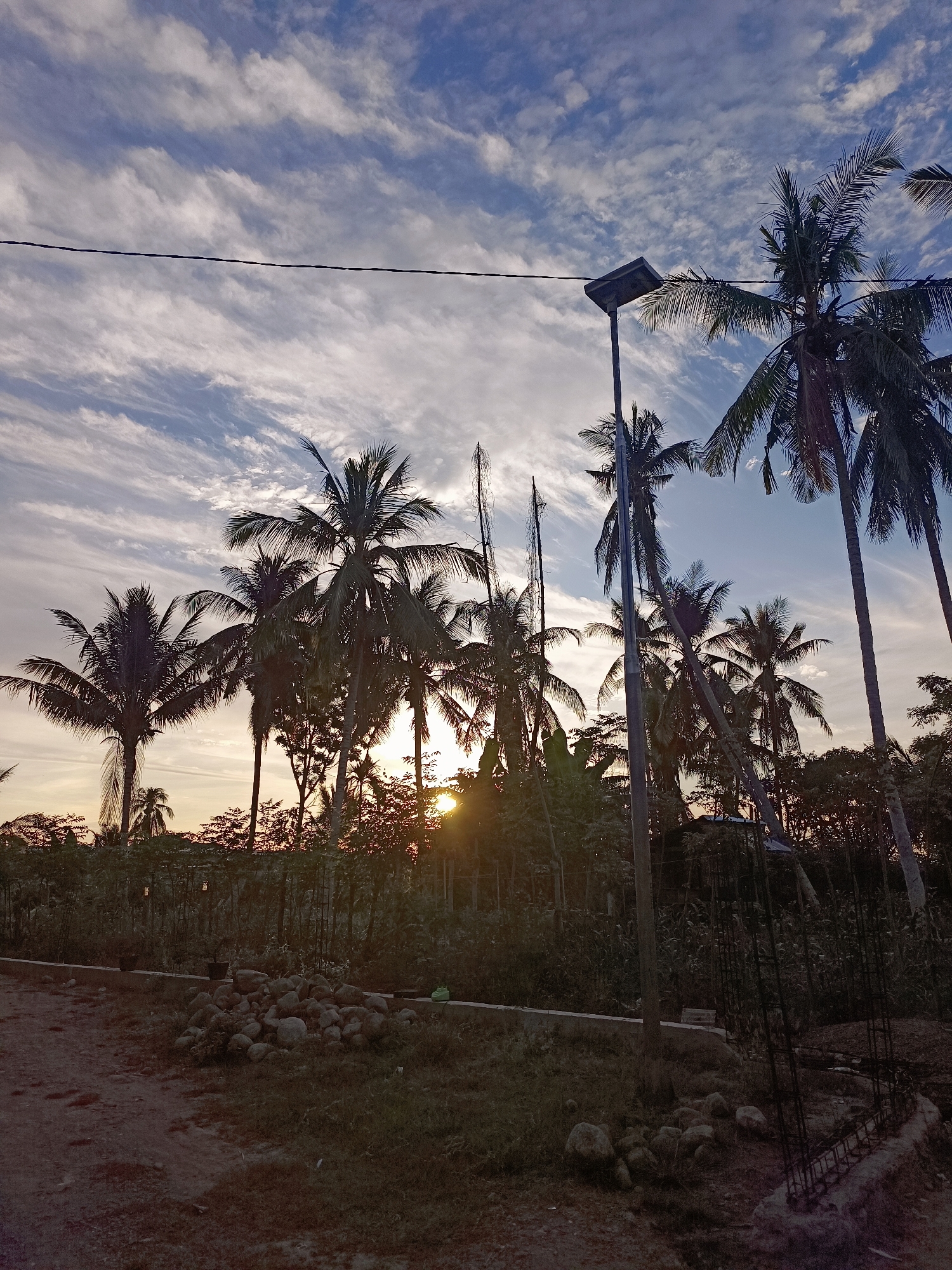 A low-angle, vertical shot captures a tropical sunset in Meunasah Raya, Indonesia. The sky is a gradient of blue and white, with wispy clouds streaking horizontally across the frame. A telephone wire stretches across the upper portion of the image. In the foreground, a dirt path leads to a grassy area scattered with rocks. A low wall runs along the edge of the grassy area, behind which a dense collection of trees and palm trees are silhouetted against the setting sun. The sun is visible as a bright orb peeking through the trees, casting a warm glow. A solar-powered street light stands to the right of the frame, its dark metal pole extending upwards towards the sky. The scene appears to be a rural or semi-rural area, possibly on the outskirts of a village. There are no people visible in the image.