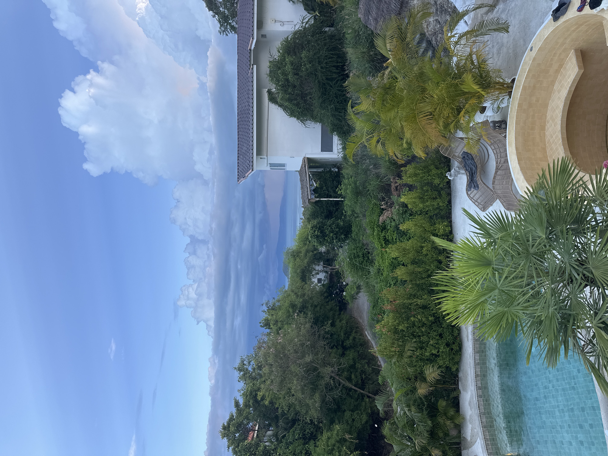 This media file depicts a tropical outdoor scene in Koh Phangan, Thailand. The foreground features a resort-style setting with a curved swimming pool with blue tiled water. Around the pool, there are lounge chairs and lush green palm trees, some with yellowing fronds, suggesting a warm climate. The area is paved with light-colored concrete.
Further back, a white building with a tiled roof is visible, partially obscured by dense green foliage and trees. A pathway winds through the vegetation. In the distance, a body of water, likely the ocean, can be seen with a hazy mountainous landscape beyond.
The sky is a prominent feature, with a bright blue expanse on the left, transitioning to dramatic, dark storm clouds on the right, indicating changing weather conditions. The lighting suggests it is either early morning or late afternoon, with the sun creating highlights on the clouds. No people are visible in the scene, and no text is present. The overall impression is one of a tranquil, yet dynamic, tropical environment.