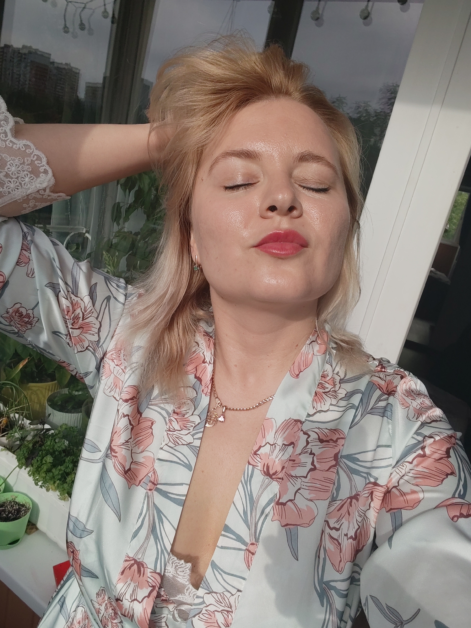 A fair-skinned blonde woman with closed eyes, glossy red lips, and a rose gold necklace poses in a floral robe. Her right arm is raised, hand resting on her head, revealing a lace sleeve. The backdrop features a window reflecting buildings, potentially the Izhevsk skyline, and greenery suggests an indoor garden. Sunlight illuminates her face.