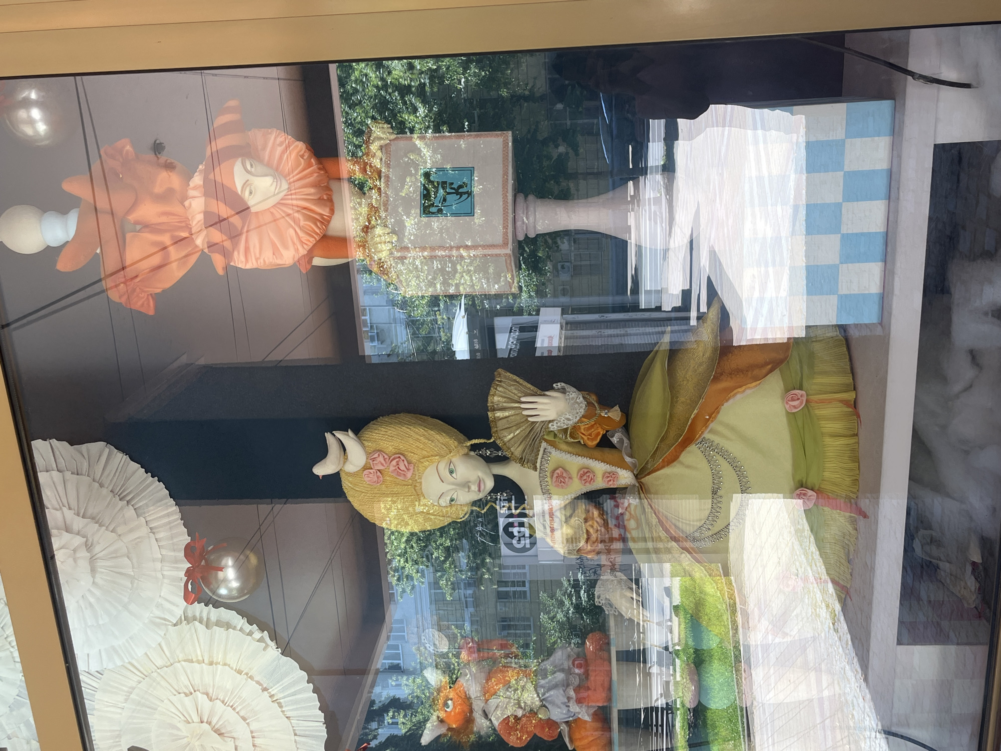 This image displays a window display, likely from a shop or exhibition, featuring elaborate mannequins and decorative elements. The primary subjects are two figures: a jester-like character with an orange ruffled collar and a large, elaborate doll dressed in a historical-style gown with a powdered wig. The jester figure has a pale face with delicately painted features and is positioned in the upper left portion of the frame. The larger doll is in the lower right, depicted from the waist up, holding a fan and adorned with pink rosettes on her wig and dress. The overall scene is set against a backdrop that includes reflections of trees and buildings, suggesting an outdoor setting visible through the window.
Notable details include the textures and materials of the mannequins' attire – flowing fabrics, intricate beadwork on the doll's dress, and ruffled elements. Several decorative objects are present, including large, white, fan-like structures in the lower left, and a metallic sphere with a red ribbon tied around it. There's also a blue and white checkered cube and a pedestal with a framed picture, possibly a book or a plaque, displaying an emblem or logo. The lighting suggests it's daytime, with natural light casting reflections on the glass. The presence of buildings and lush greenery in the reflections provides location cues.
No discernible text is clearly visible and legible within the display itself. However, some blurry text can be seen in the reflections on the window, but it's too indistinct to read. The overall impression is one of a theatrical or whimsical presentation, potentially related to a historical theme or a special event. The image is a vertical shot, taken at a slight angle.