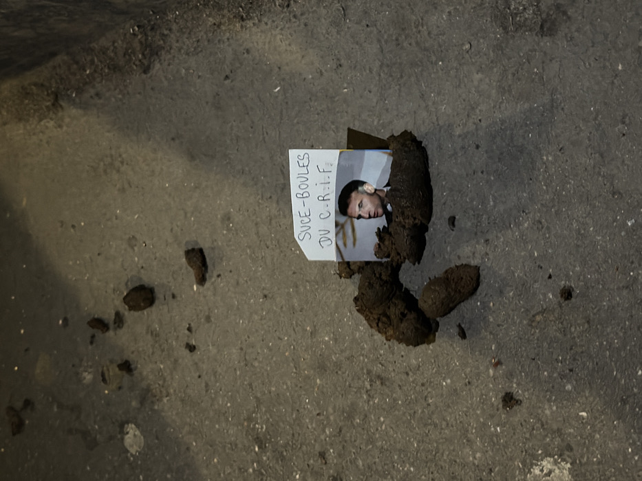 The scene depicts a derogatory display on a dark grey urban pavement in Paris, France. Centered in the frame is a cluster of dark brown organic matter, consistent with animal feces, arranged in several distinct masses. Inserted vertically into this matter are two items: a color photographic print and a handwritten note. The photograph shows a headshot of French President Emmanuel Macron, facing towards the right side of the frame. Directly above and partially overlapping the photograph is a small piece of white paper, on which "SUCE-BOULES DU C-R-I-F" is handwritten in dark ink. Minor debris, including small dark particles and specks of dirt, is scattered across the pavement surrounding the central arrangement.