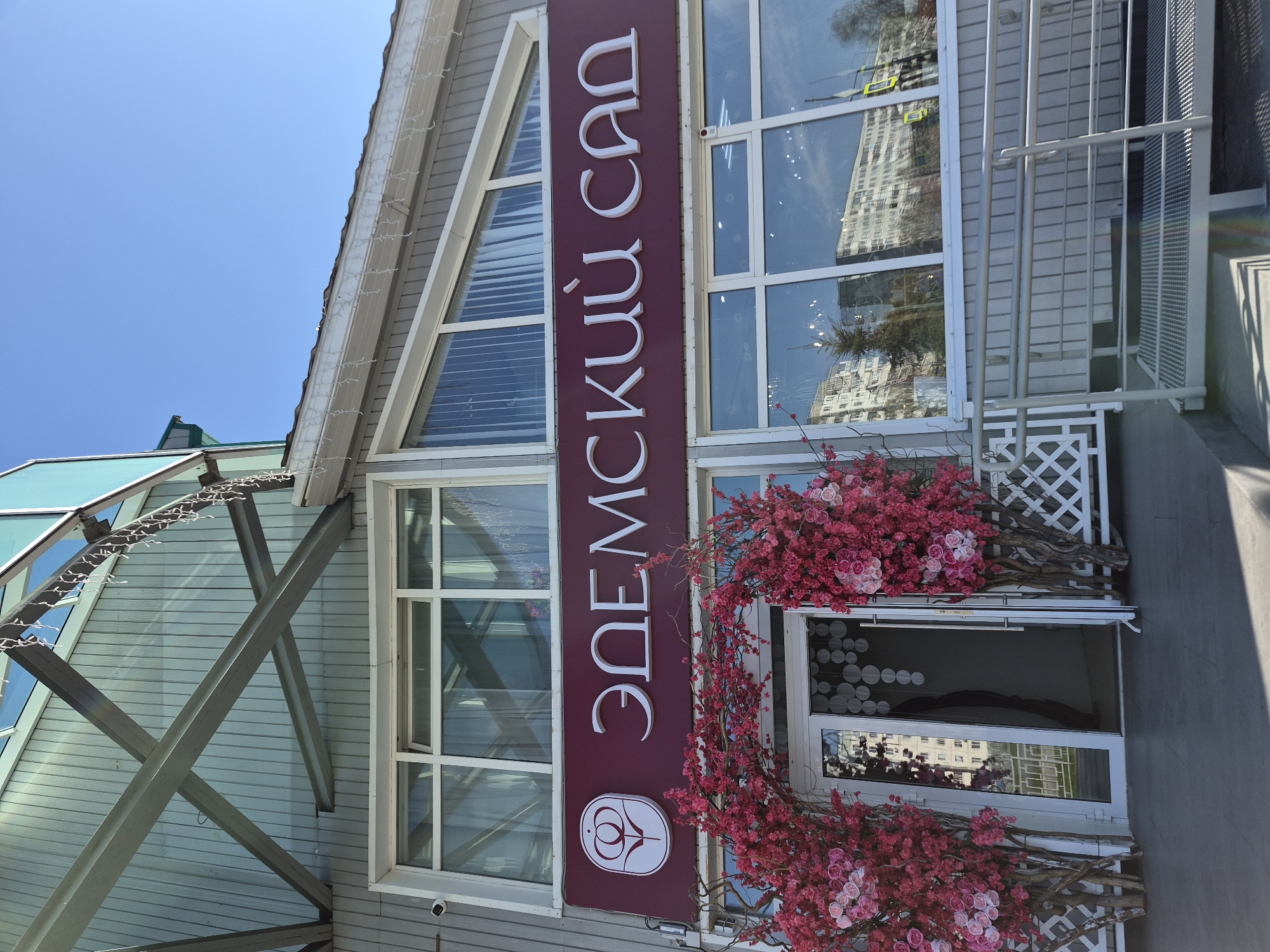 The image captures the entrance to a building with a sign in Russian that says "Eden Garden" (ЭДЕМСКИЙ САД). The building has large windows, and the entrance is decorated with pink floral arrangements. Reflections of nearby buildings are visible in the windows. The building appears to be in the city of Tobolsk, Russia. The scene is outdoors during the day.