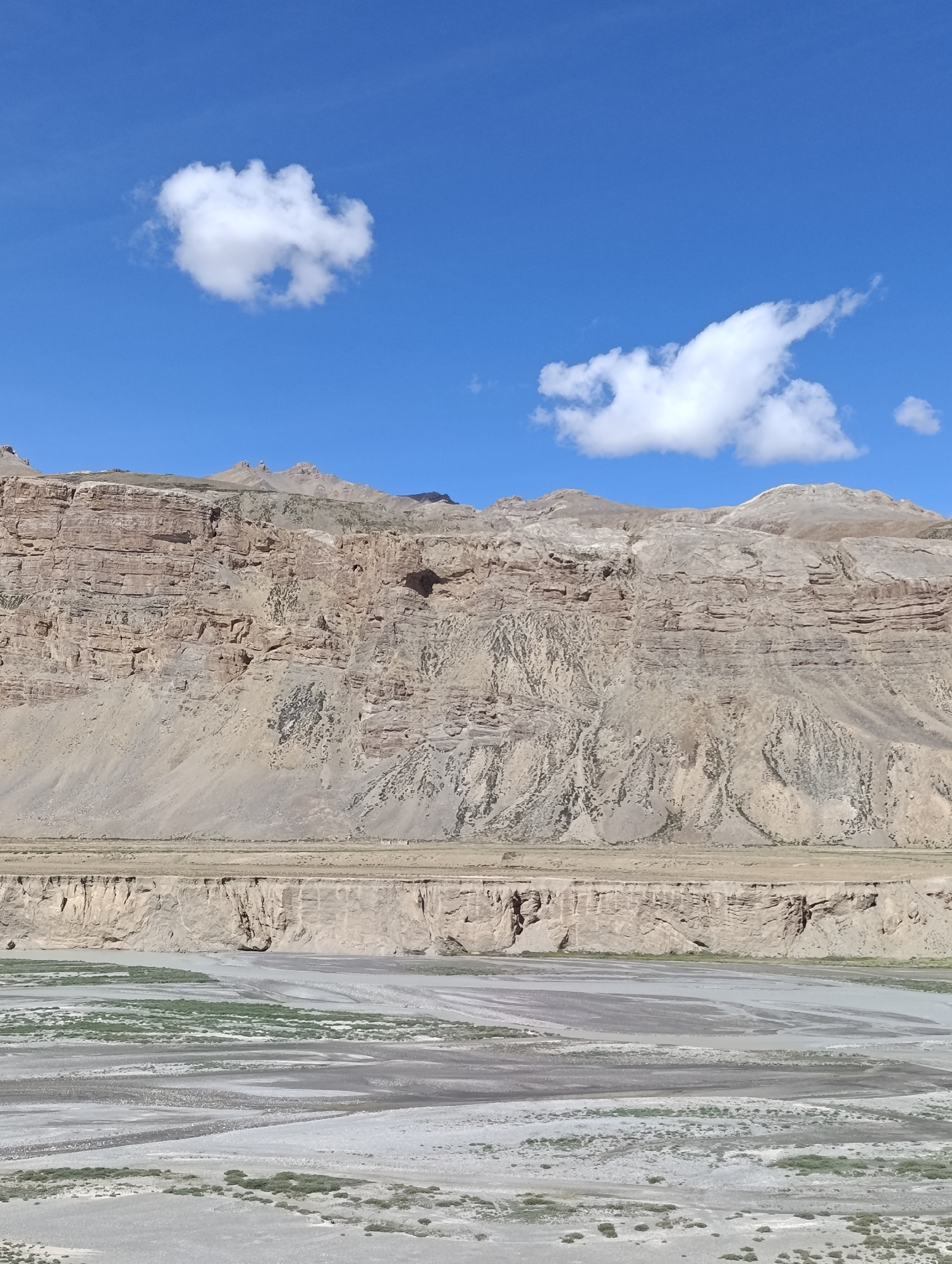 A vertical, full shot captures a vast, arid mountain landscape under a bright blue sky dotted with fluffy white clouds. The scene is divided into three horizontal bands: the sky, the mountains, and the riverbed. The sky occupies the top third of the image, a clear, vibrant blue. Two distinct clouds are visible: a smaller, more amorphous one on the left and a larger, elongated one on the right.

The middle section is dominated by a massive, layered mountain range. The rock faces are a mixture of tan, brown, and grey, exhibiting clear stratification and erosion patterns. There are visible crevices and shadows that suggest depth and ruggedness. The mountain's base transitions into a flat, dry plain.

The bottom third of the image depicts a wide, shallow riverbed. The ground is a mix of sand, gravel, and some sparse, green vegetation. The water appears to be low, forming sinuous channels and pools across the broad expanse. The overall impression is one of a remote, natural environment, likely in a high-altitude desert region. The specific location is unknown, but the geological features suggest a mountainous area of India.