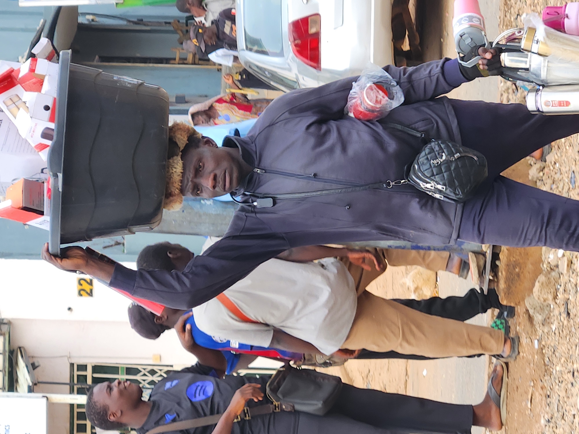 A man in a dark blue zip-up jacket and dark pants carries a large black basin filled with cardboard boxes on his head. He is walking towards the right of the frame, with his left arm raised to balance the basin. He has a small black quilted bag slung across his chest. In his right hand, he is carrying a pink water bottle and a metal container with a handle, as well as other items wrapped in plastic. Another man in a dark blue shirt with a Spotify logo is to the left, wearing a dark shoulder bag. Behind him, another person wears a light gray t-shirt and tan pants. In the background, a silver car is parked to the right, and a building with the number 22 on it is visible. The ground is covered in dirt and small rocks.