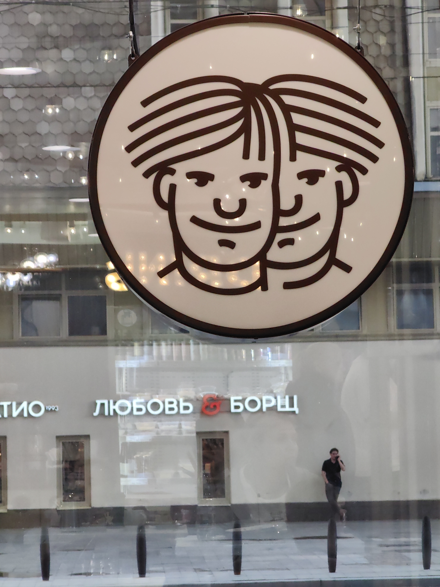 A circular sign with a brown outline and a white interior is hanging in front of a building. The sign depicts two cartoon faces with brown stripes for hair. Below the sign, the building has the words "ЛЮБОВЬ & БОРЩ" and "1993" in white lettering. A person in a black shirt and grey pants is standing and talking on a cell phone in front of the building. The environment appears to be urban, with windows and reflections visible in the background.