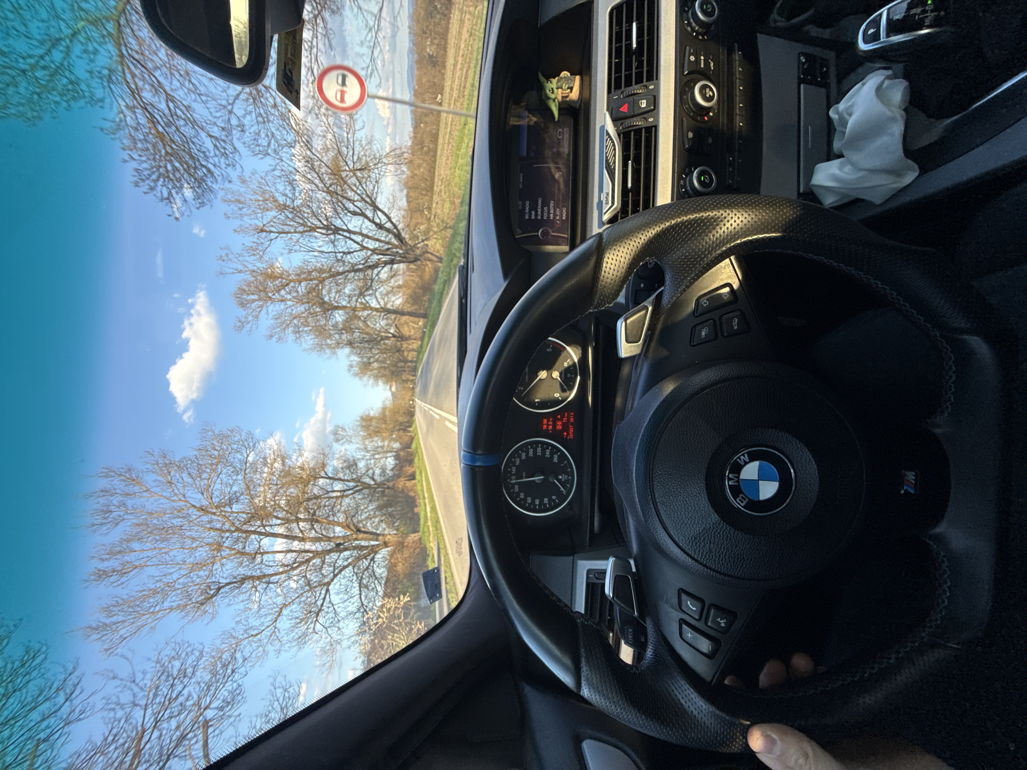 The image is a first-person perspective from inside a BMW car, presumably on a road trip in Gorna Rositsa, Bulgaria. The steering wheel, with the BMW logo and "M" badge, dominates the foreground. The dashboard is visible, featuring a screen displaying radio stations and car information (such as time, temperature, and odometer reading). A small Yoda figurine sits on the dashboard. Through the windshield, a road stretches into the distance, flanked by bare trees against a blue sky with some clouds. A road sign is also visible. A partially visible hand is gripping the steering wheel. A white cloth rests on the center console.