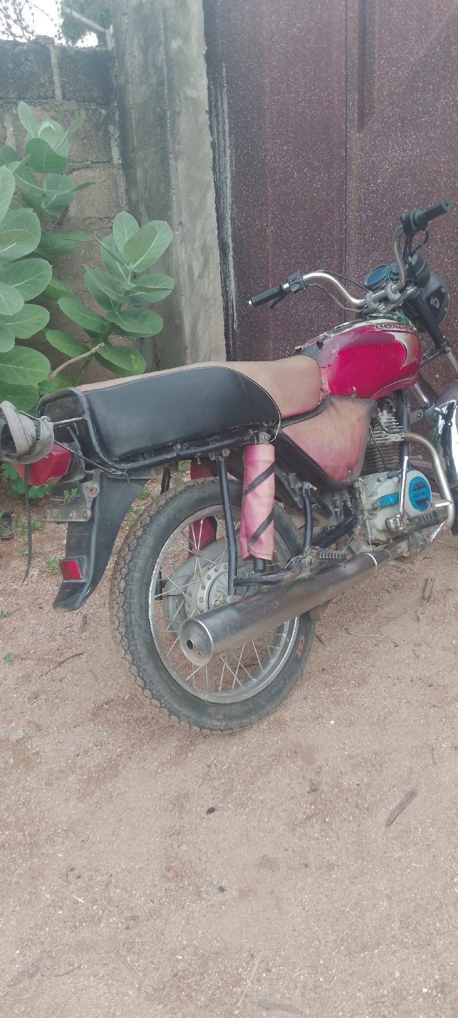 A red and black motorcycle is parked on a dirt surface, with a textured brown metal gate and a grey concrete wall in the background. The motorcycle has a worn black seat, a red fuel tank, and a silver exhaust pipe. There are green plants to the left of the motorcycle, and the ground is covered in sand and small stones. The scene appears to be outdoors, possibly in a residential or storage area in Jalingo, Nigeria.