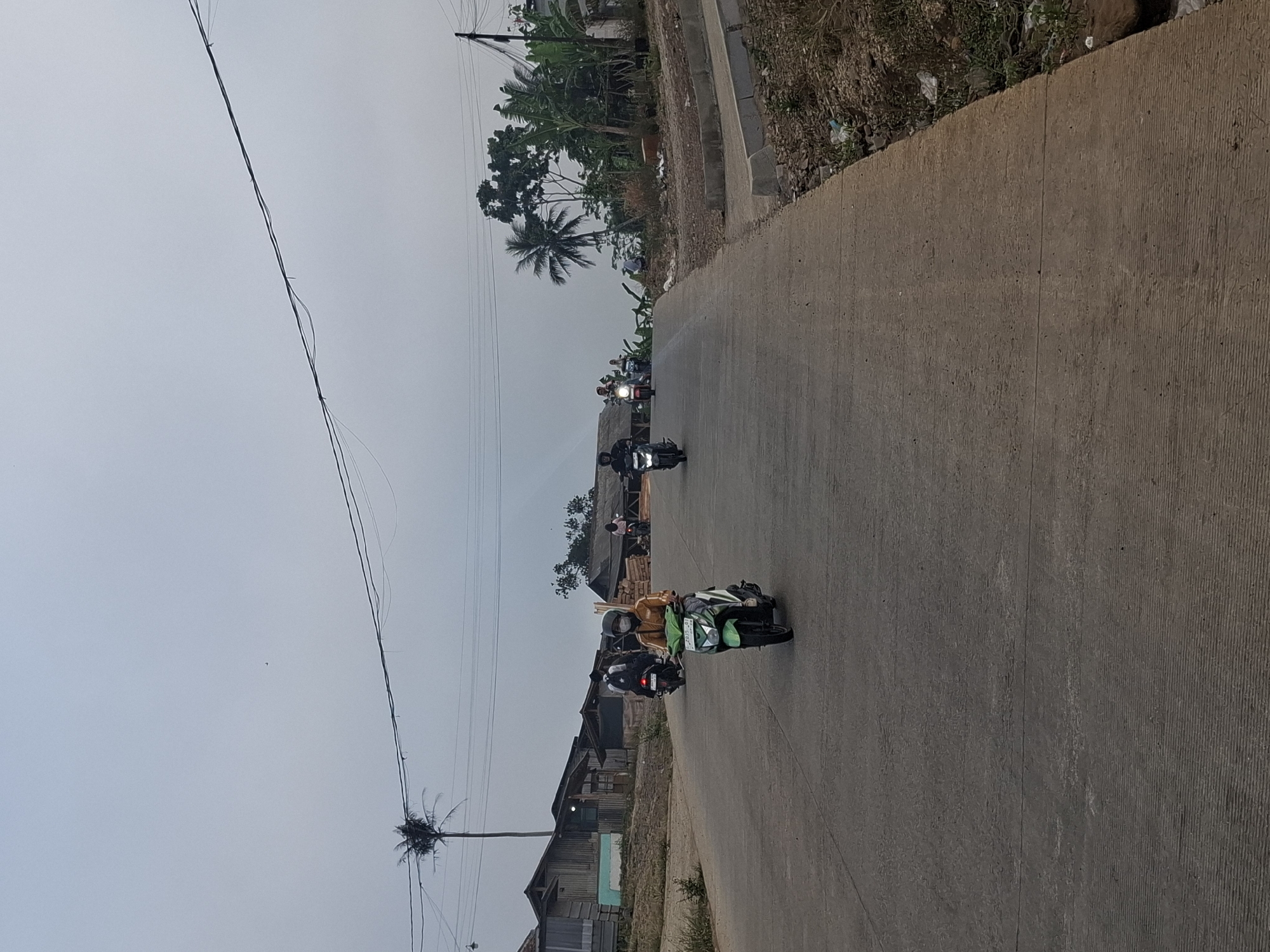 The image depicts a paved, uphill road in what appears to be a semi-rural or suburban area of Ciheulang Tonggoh, Indonesia. The sky is overcast, suggesting either early morning or late afternoon. Several individuals on motorcycles are traveling up the road. In the foreground, a person wearing a yellow jacket and a face mask is riding a green motorcycle. Behind them, other motorcyclists are visible, some carrying passengers. The road is bordered by a dry, somewhat overgrown shoulder with debris. On the left side of the road, several simple, weathered structures with tin roofs are visible, some appearing to be houses or workshops. Palm trees and other tropical vegetation are present in the background, contributing to the Indonesian setting. Power lines are strung across the sky.