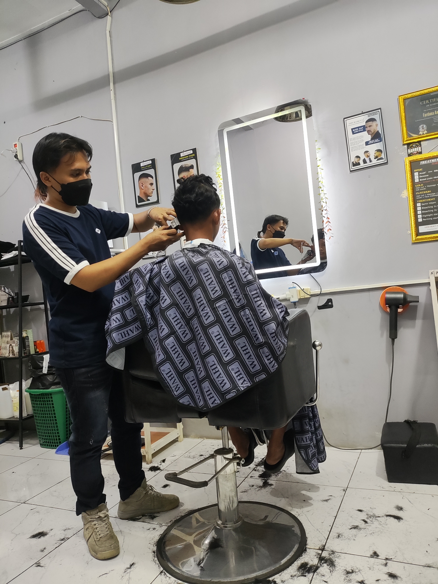 In a barbershop located in Wanajaya, Indonesia, a male barber, wearing a dark blue t-shirt and black face mask, is giving a haircut to a client. The client is seated in a black barber chair, draped with a cape featuring a repeating "WAHL" logo pattern. The barber is holding electric clippers in his right hand, actively cutting the client's hair. The environment appears to be a typical barbershop with a large illuminated mirror behind the barber, reflecting the barber and client. On the wall, there are framed certificates and posters displaying different hairstyles. To the left, shelves are stocked with various hair care products. The floor is tiled and shows scattered hair clippings, indicating recent activity. An orange hair dryer is mounted on the wall to the right.