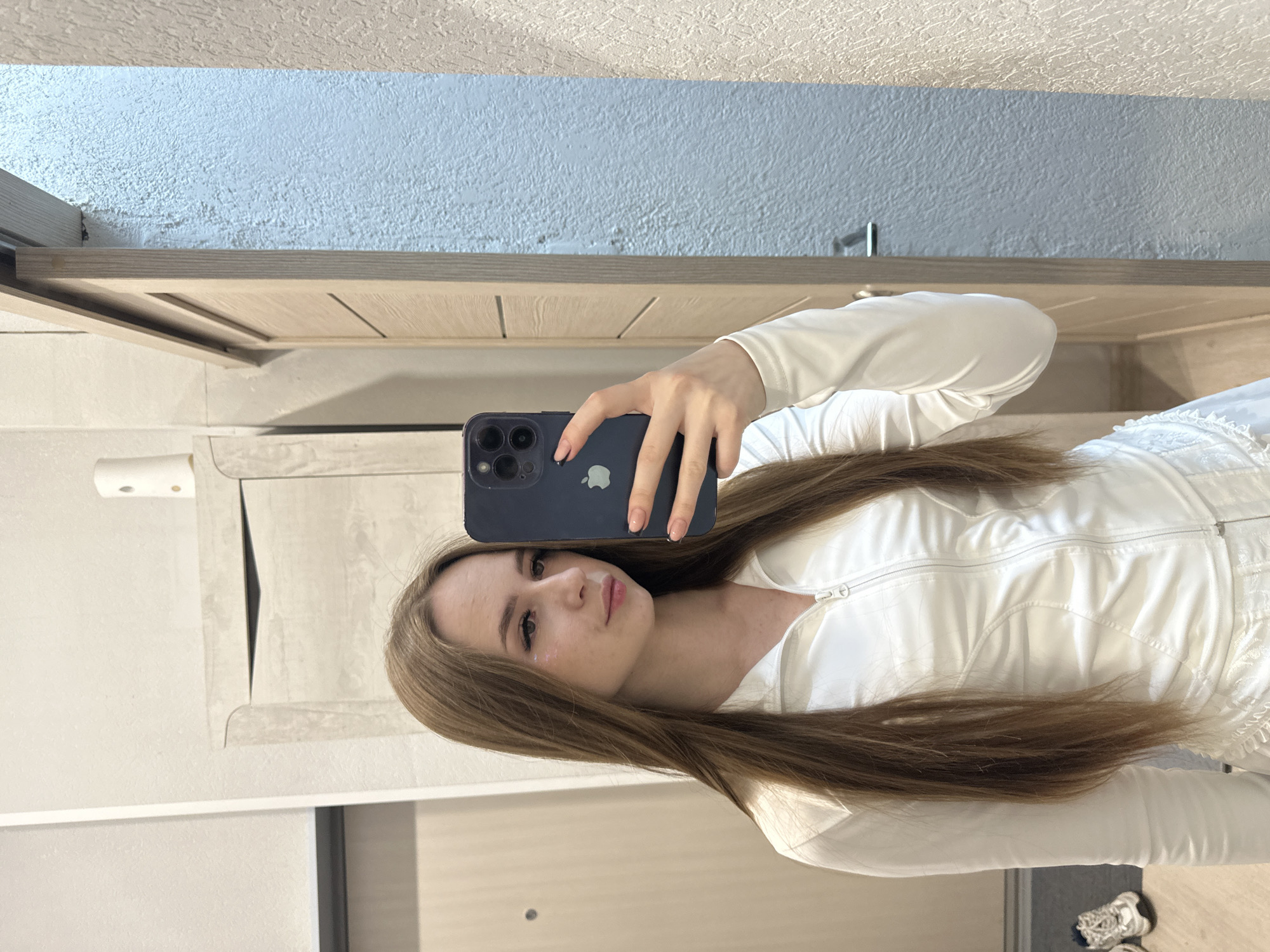 A young woman is taking a selfie in an indoor setting. She is holding a blue smartphone in her right hand, covering part of her face. She has long, straight brown hair. She is wearing a white zippered top. The background includes a textured light blue wall and a beige door or cabinet. There is a partial view of what appears to be a wall-mounted paper towel dispenser and another door or panel.