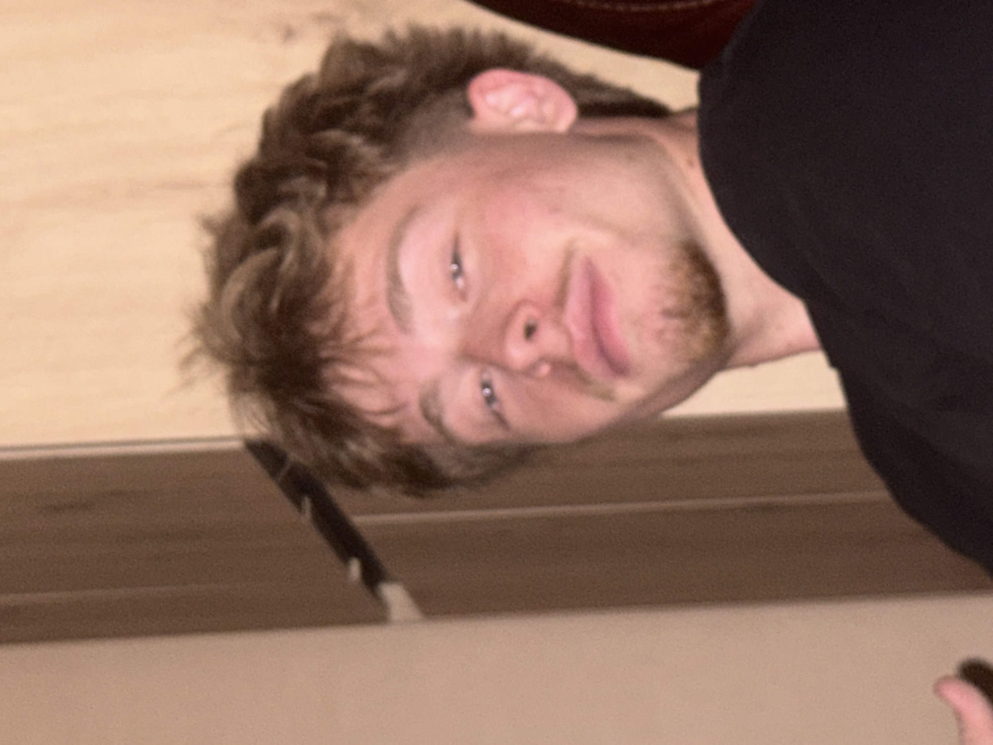 The image displays a close-up portrait of a young Caucasian male, appearing to be in his late teens or early twenties, situated indoors in Sofia, Bulgaria. The subject possesses short, textured brown curly hair, styled with volume on top and closely trimmed sides. He has light brown facial hair, comprising a mustache and a small goatee on his chin. His eyes are light-colored, and he gazes directly towards the viewer with a subtle smirk, characterized by slightly puckered lips. He is dressed in a black, short-sleeved, crew-neck top. The background on the left consists of a plain, light beige-colored wall. To the right and behind the subject, a vertical dark brown wooden panel or doorframe is visible, exhibiting wood grain texture. Warm, diffused lighting illuminates the scene. A dark, possibly upholstered object, likely furniture, is partially visible behind his right ear. A small portion of his left hand is seen at the lower left edge of the frame.