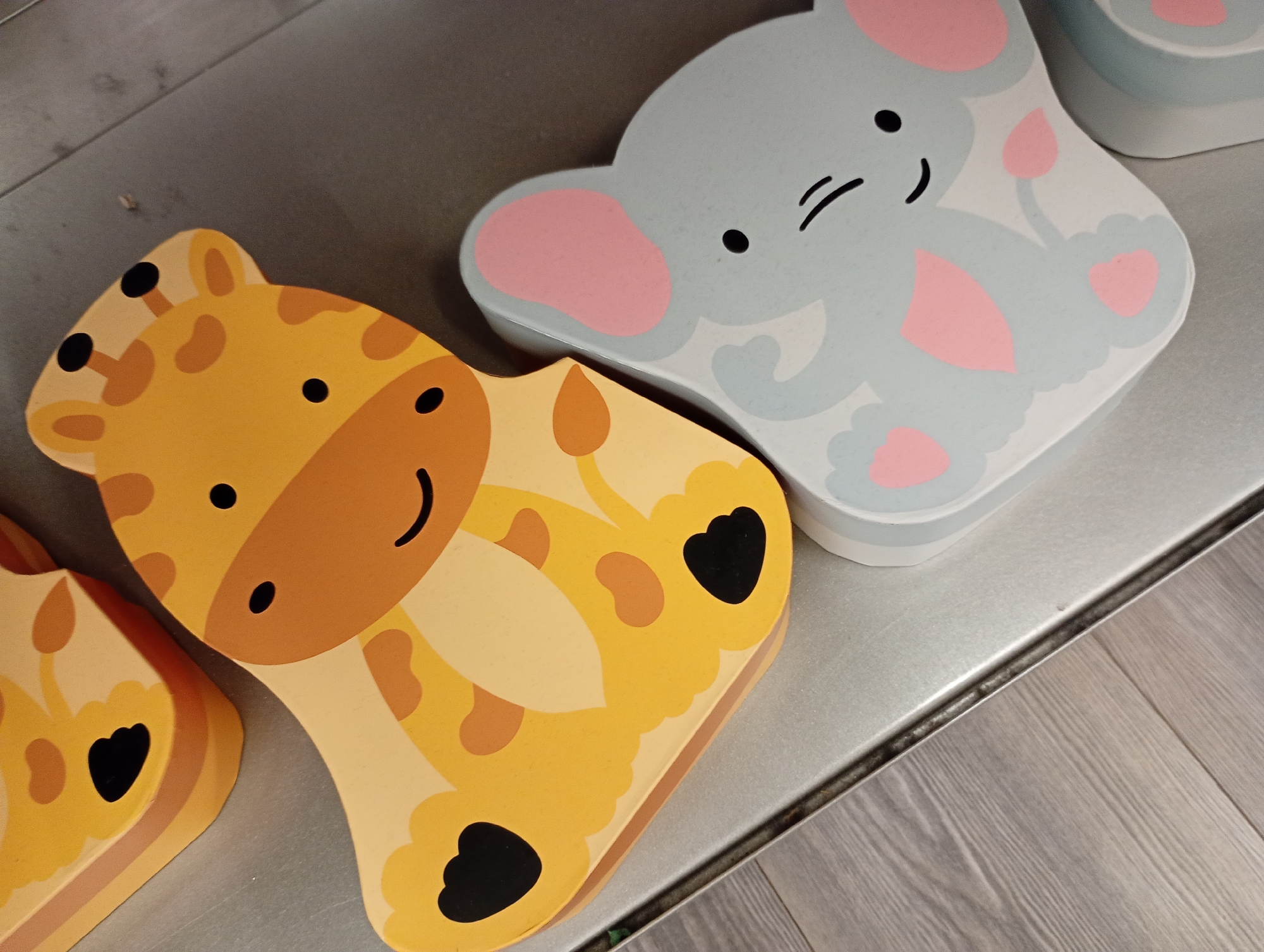 The image shows two cartoon animal-shaped containers, a giraffe and an elephant, placed on a metallic shelf. The giraffe container is yellow with brown spots and depicts the animal's head and neck. The elephant container is light blue with pink accents, showing the elephant's face and body. The containers are positioned next to each other, with the giraffe on the left and the elephant on the right. The background consists of a grey wooden floor. The scene is indoors, likely in a retail environment or a home. The image does not contain any information about Bad Lauterberg im Harz, Germany.