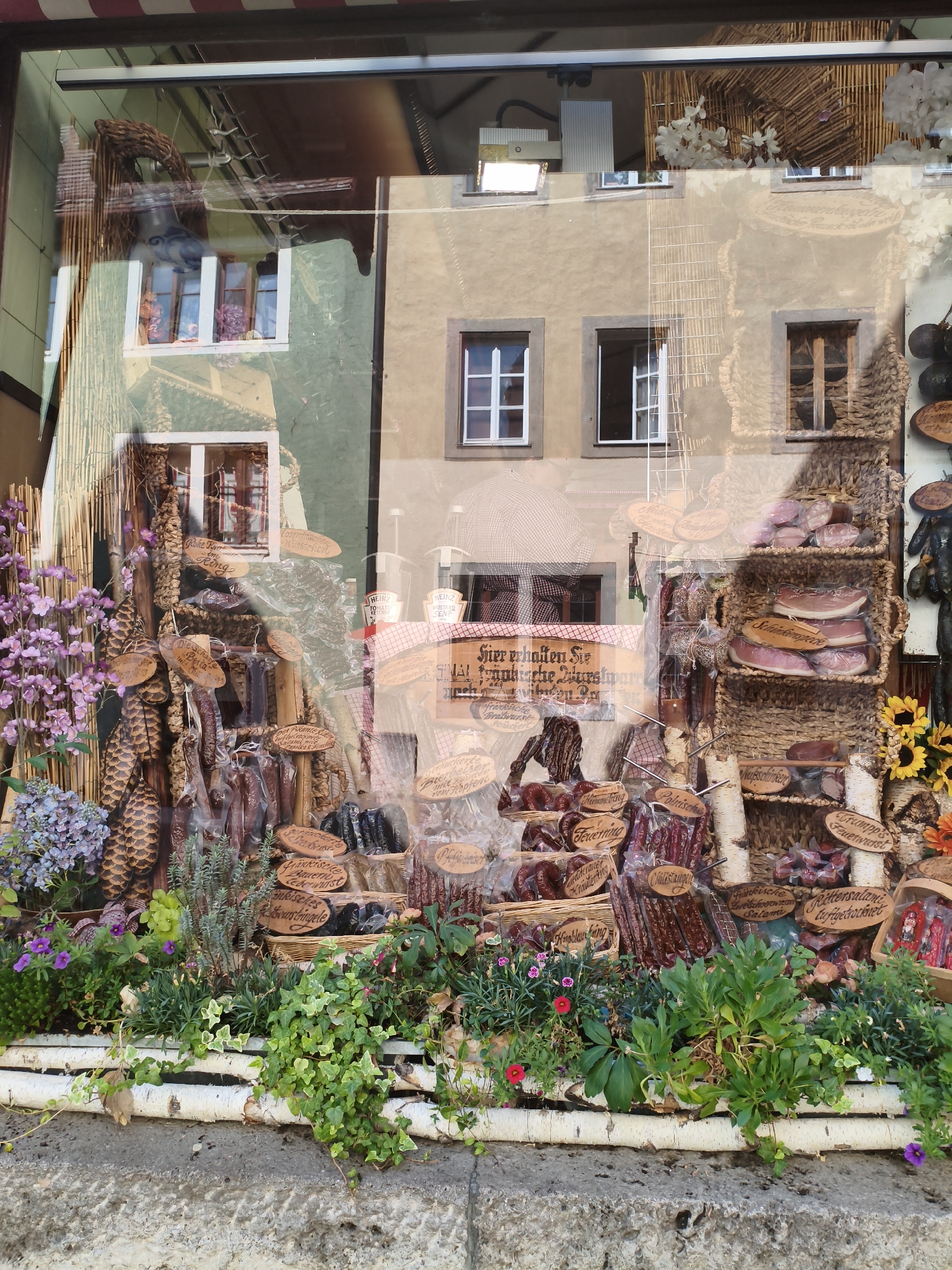 The image shows a reflection in a shop window in Rothenburg ob der Tauber, Germany. The reflection displays a facade of a building with several windows. In the foreground of the reflection is a display of cured meats and other food products arranged in baskets and hung from ropes. These products appear to be artisanal and traditional, with wooden signs labeling them in German. There are also decorative elements such as pine cones, branches, and artificial flowers. The overall impression is a traditional German butcher or delicatessen shop. The real scene outside the shop window shows greenery, including flowers and ivy, along the bottom of the frame, and a stone wall.