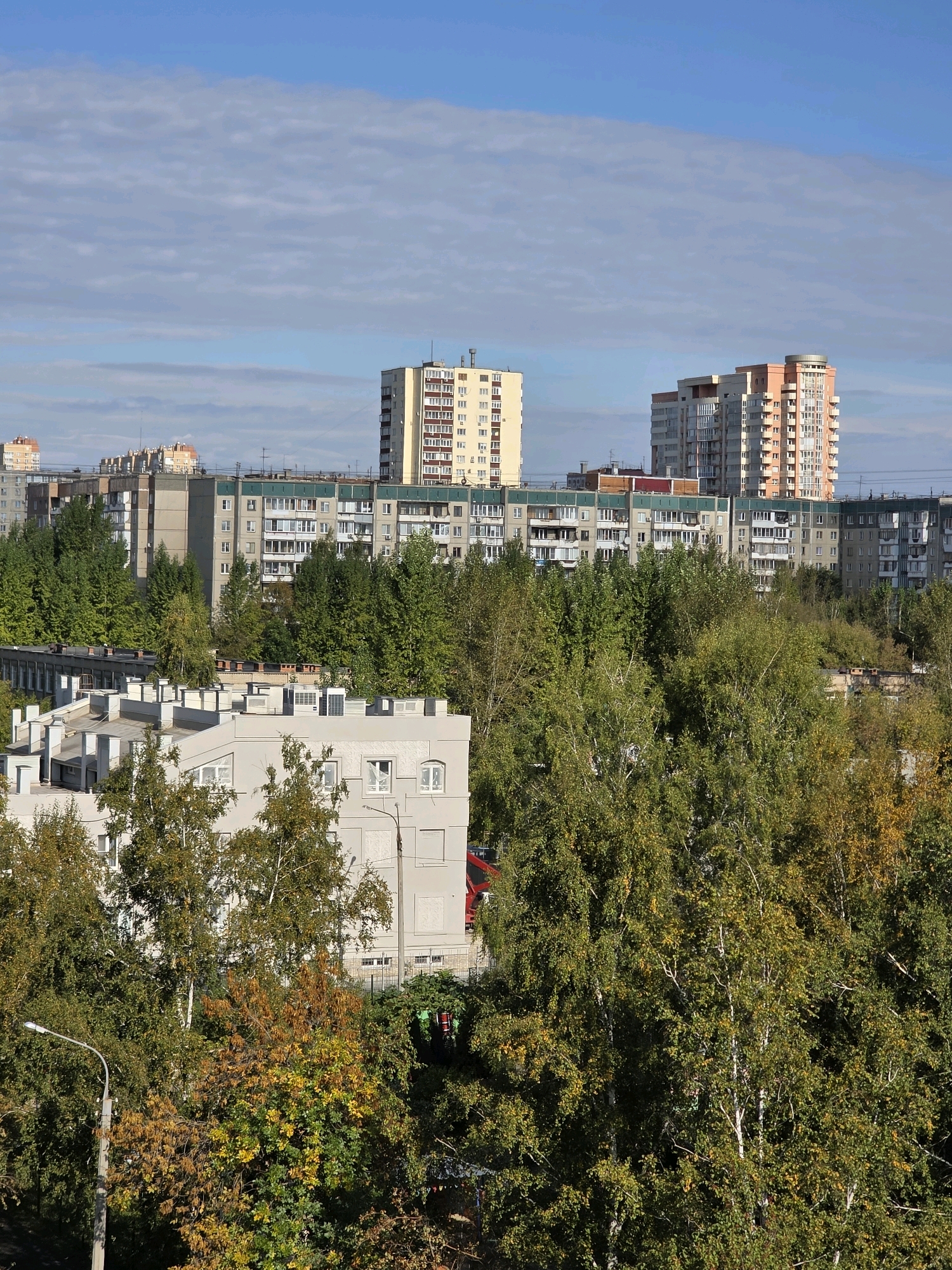 This is an aerial photograph of a residential area in Chelyabinsk, Russia, captured on a sunny day with a partly cloudy sky. The scene is dominated by multi-story apartment buildings and a dense canopy of trees, many of which are beginning to show autumn colors with yellow and orange foliage mixed with green.
In the foreground, a light-colored apartment building with multiple windows and what appear to be air conditioning units on its roof is partially obscured by trees. To the left, a street lamp is visible.
Behind the foreground building, a row of longer, mid-rise apartment buildings with many windows stretches across the frame. These buildings are of varying architectural styles and colors, with some featuring green accents.
Further in the background, two taller apartment buildings stand out. The one on the left is a cream-colored building with a grid-like pattern of windows. The building on the right is taller and more modern, with a mix of white, orange, and beige facades and a rounded section with glass.
The environment appears to be a typical urban setting in Russia, with a mix of older and newer residential architecture surrounded by significant green spaces. The presence of deciduous trees suggests the photograph was taken in the fall. There are no people or vehicles clearly visible in the frame. The overall impression is of a tranquil, daytime urban landscape.