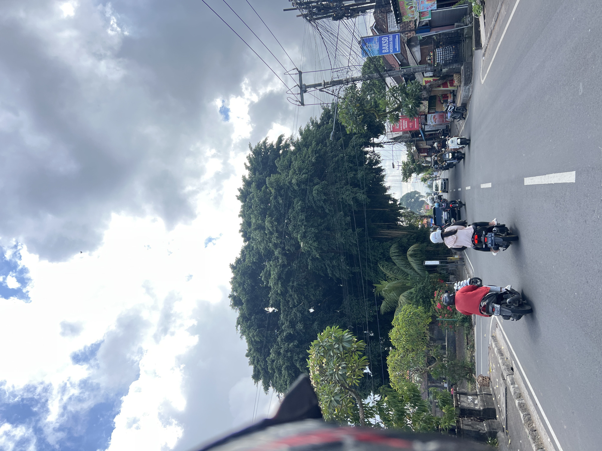 This image depicts a street scene in Abiansemal, Indonesia. The perspective is from a moving vehicle, likely a scooter, as evidenced by the partial view of a helmet on the left side of the frame. The road is paved and has white lane markings. Several scooters are visible on the road, with riders wearing helmets. In the foreground, two scooters are traveling away from the viewer. The scooter on the left is ridden by a person in a red shirt, and the scooter on the right is ridden by a person in a light-colored hoodie.
The environment is lush and tropical, with large trees dominating the landscape, particularly a massive, dark green tree with dense foliage behind the scooters. Smaller trees and plants are also present along the roadside. Buildings with signs and utility poles with a complex network of wires line the right side of the road, suggesting a commercial or residential area. One prominent sign reads "BAKSO MERDEKA SOLO." The sky is partly cloudy, with a mix of bright white clouds and darker, overcast areas. The overall impression is that of a typical day on a busy street in a tropical urban or semi-urban setting.