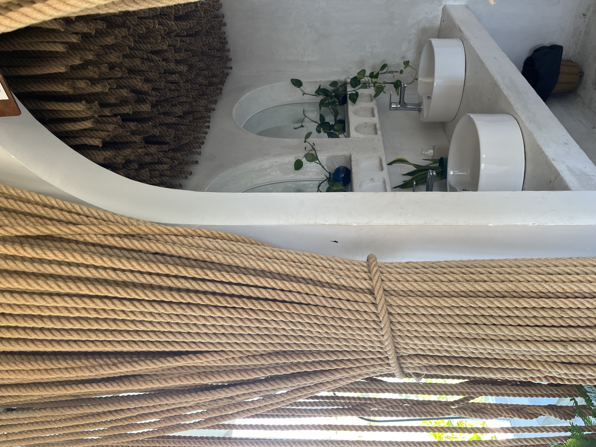 This media file captures a uniquely designed interior space, likely a washroom or vanity area, characterized by its rustic and natural aesthetic. In the immediate foreground, numerous thick, light brown ropes are strung horizontally, creating a textured screen or barrier. These ropes appear to be jute or sisal, with one section tightly bound vertically by another rope, forming a decorative and possibly structural element.
The background reveals a white, possibly plastered or cement, wall and fixture. To the left, a textured wall section is covered with tightly packed, shorter segments of darker brown rope, complementing the foreground ropes and adding depth. To the right, two circular mirrors are set into arched alcoves within the white structure. Trailing green vining plants, possibly Pothos, are decoratively placed around the mirrors and along the white surface. Below these, two white, ceramic, bowl-shaped sinks are mounted, each equipped with a silver faucet. A pump bottle (likely soap) and a snake plant are visible near the lower sink. Further to the right, a dark object, possibly a bag, rests inside a woven basket on a white ledge. A small blue decorative bottle or vase is also visible near the upper mirror.
The scene is brightly lit, suggesting natural daylight, creating subtle shadows and highlights on the textured surfaces. No people are present, and no specific activity is taking place. The overall impression is one of a clean, minimalist, and organic design often found in resort or spa settings, aligning with a tranquil atmosphere in a location like Đà Nẵng, Vietnam. There is no visible text in the image.