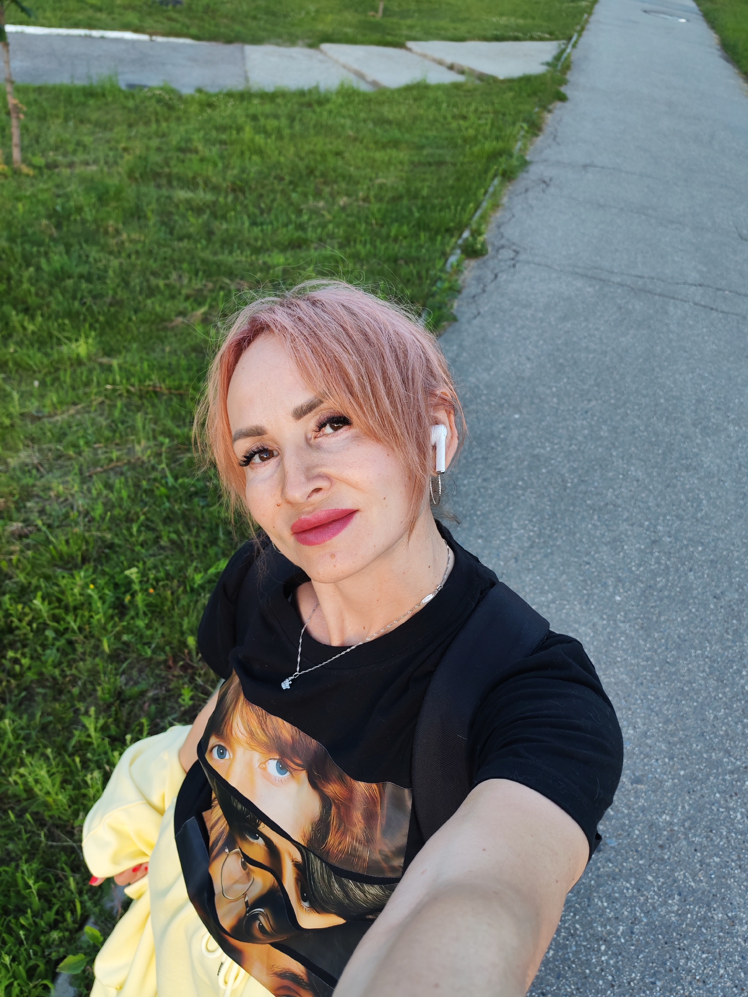 A woman with pink hair and makeup is taking a selfie outdoors. She is wearing a black t-shirt with a graphic print of eyes, a silver necklace, and white wireless earbuds. She is also carrying a yellow garment. The background shows a paved walkway and a grassy area. The image appears to have been taken during the day in a park or recreational area.
