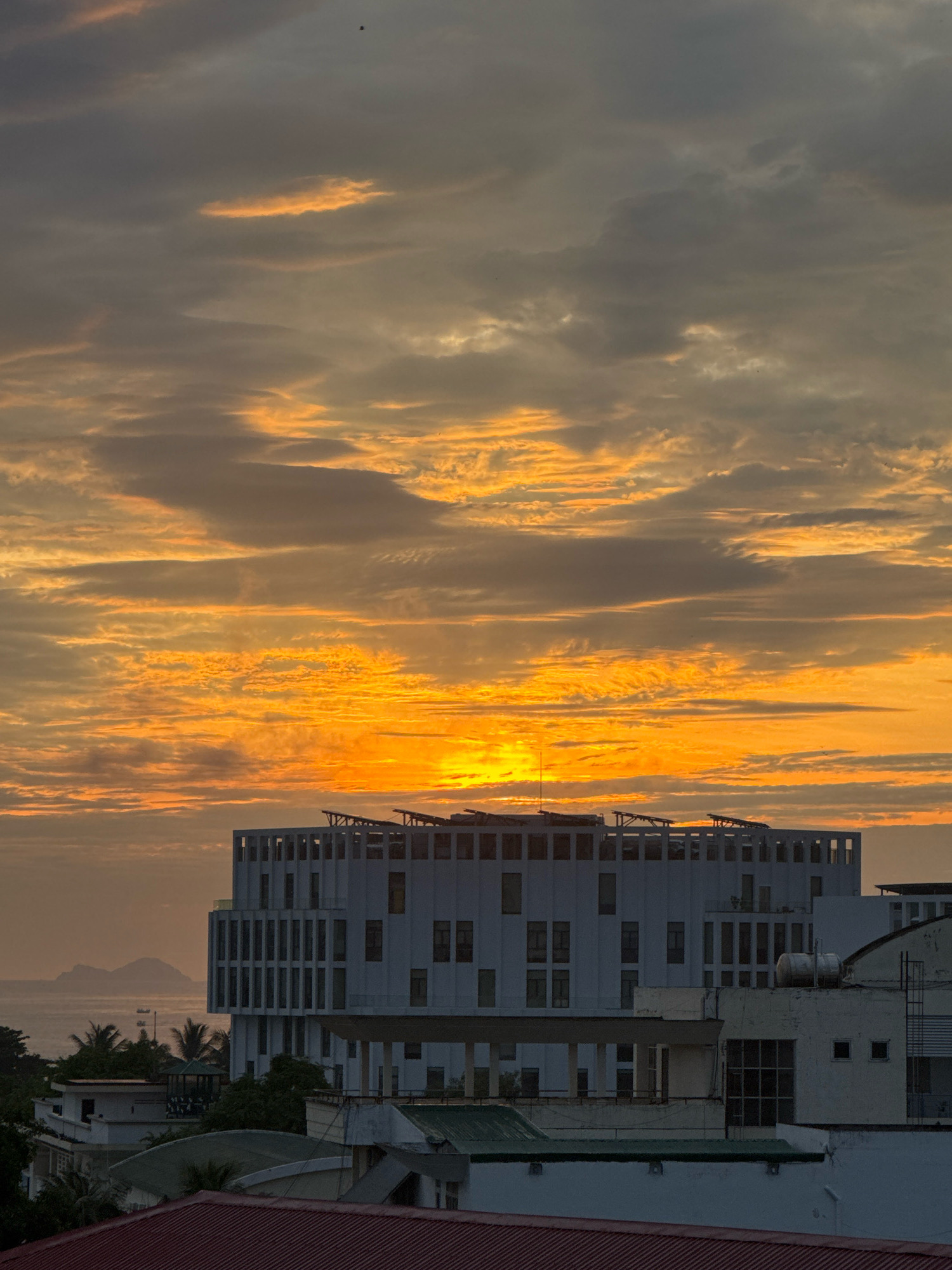 The image captures a vibrant sunset over Nha Trang, Vietnam. The sky is a canvas of fiery oranges and yellows, with scattered grey clouds adding depth and texture. The sun, a bright disc of light, is partially obscured by the clouds, casting a warm glow across the scene.
In the foreground, several buildings are visible, their architecture indicative of a coastal city. The most prominent is a modern, multi-story white building with a series of vertical window panels and what appear to be solar panels on its roof. To its right, another building with a more industrial aesthetic stands, featuring a water tank and multiple windows. In the lower foreground, parts of red-tiled roofs and green-roofed structures are visible, suggesting a mix of residential and commercial properties.
In the background, a body of water, likely the ocean, stretches towards the horizon. Distant islands or mountains can be seen as faint silhouettes against the twilight sky. A small boat is also visible on the water, adding a touch of life to the seascape. Palm trees are interspersed among the buildings, characteristic of a tropical location. A single bird is captured in flight against the vast expanse of the sky, a small detail that enhances the serene atmosphere.