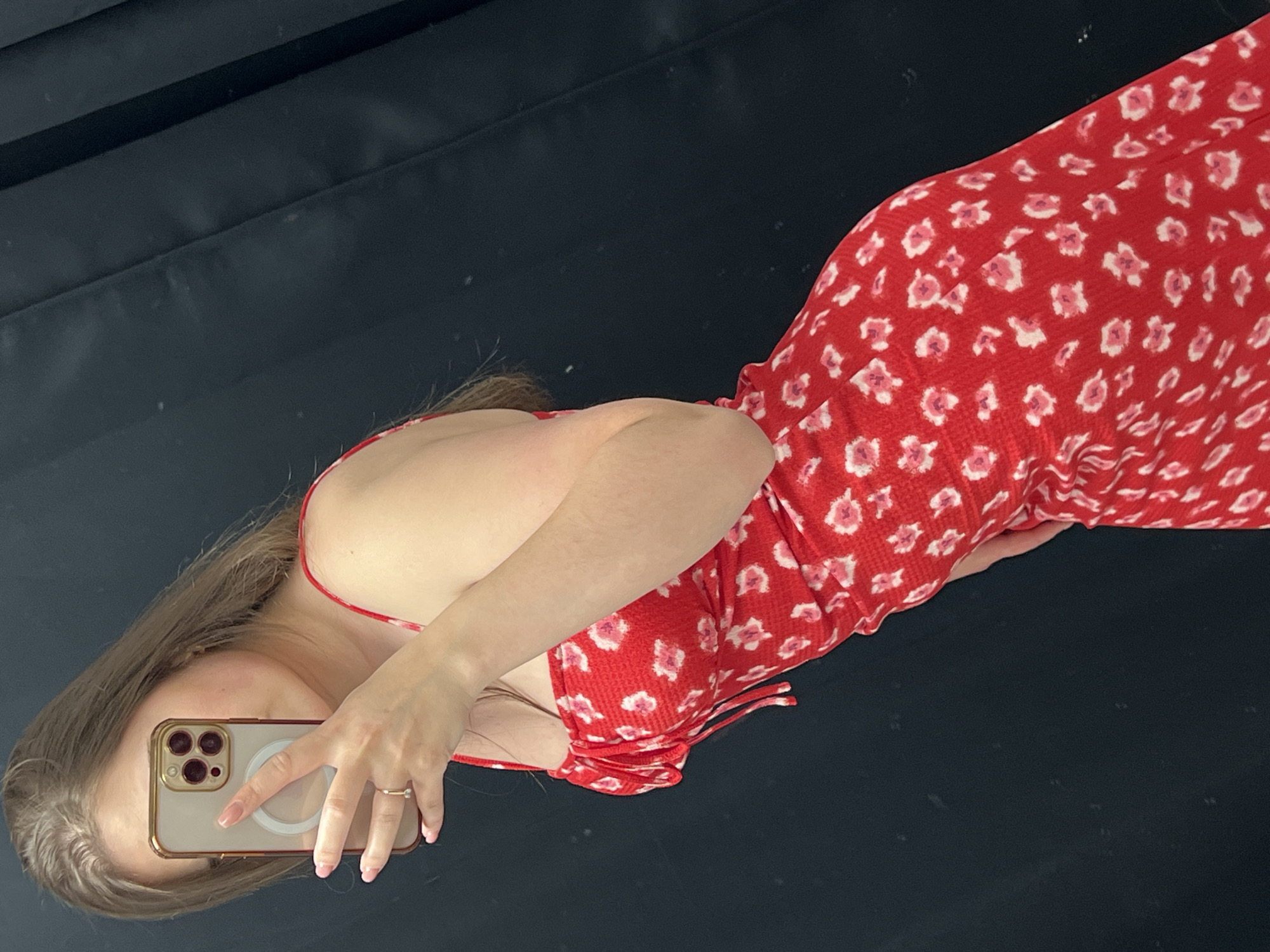 A fair-skinned person takes a mirror selfie with an iPhone 13 Pro in a gold case. The person wears a red, floral patterned dress. The person has long brown hair and manicured pink nails. The mirror's background has both a black wall and a dark, reflective surface. There is no indication of the location being in Ternopil, Ukraine.