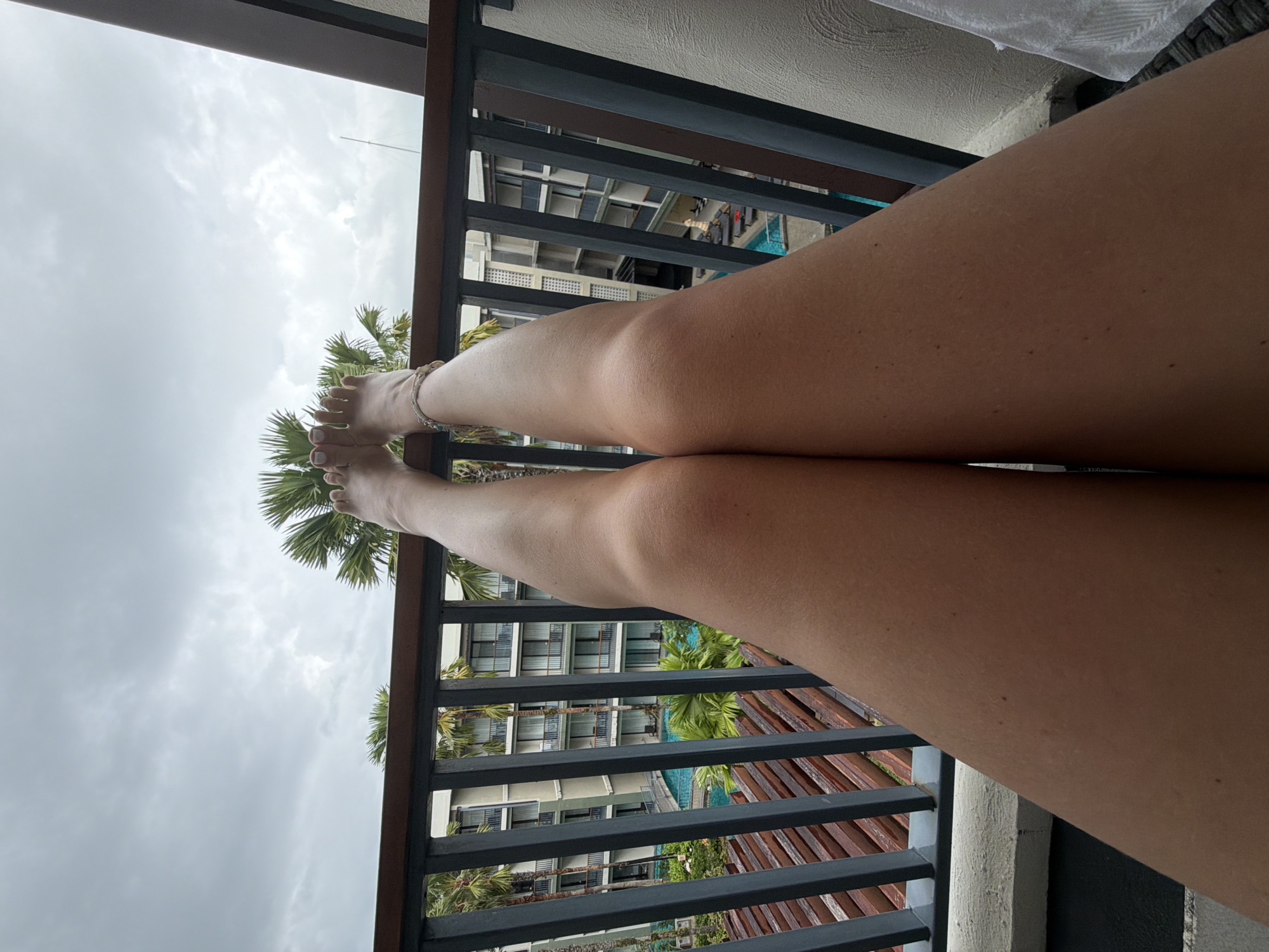 A first-person perspective shot shows a person's legs and feet resting on a balcony railing, with a resort and cloudy sky in the background. The person's legs, tanned and smooth, are positioned vertically in the frame, with their knees visible. The feet are at the top of the image, with toes pointing upwards. The person is wearing a thin anklet on their left ankle.
The balcony railing is made of dark metal bars and a wooden top. Beyond the railing, a resort building with multiple floors, balconies, and windows is visible. A swimming pool with turquoise water is situated in front of the resort. Palm trees with green fronds dot the landscape. The sky is overcast with gray clouds. The overall scene suggests a tropical resort vacation, likely in Bang Muang, Thailand, given the architecture and foliage.